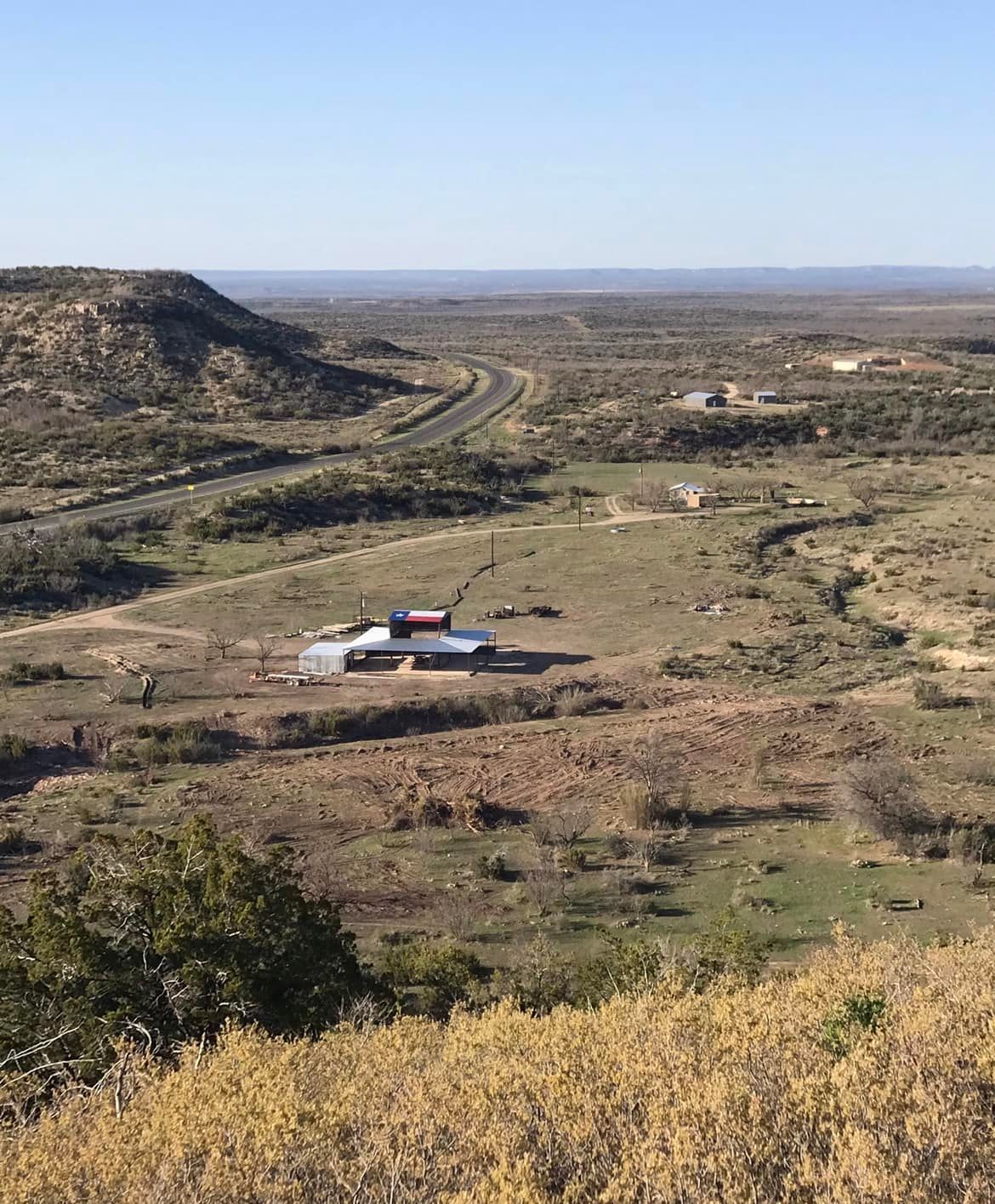 Wide dry valley with a winding road, low hills, and a small white building under a clear blue sky