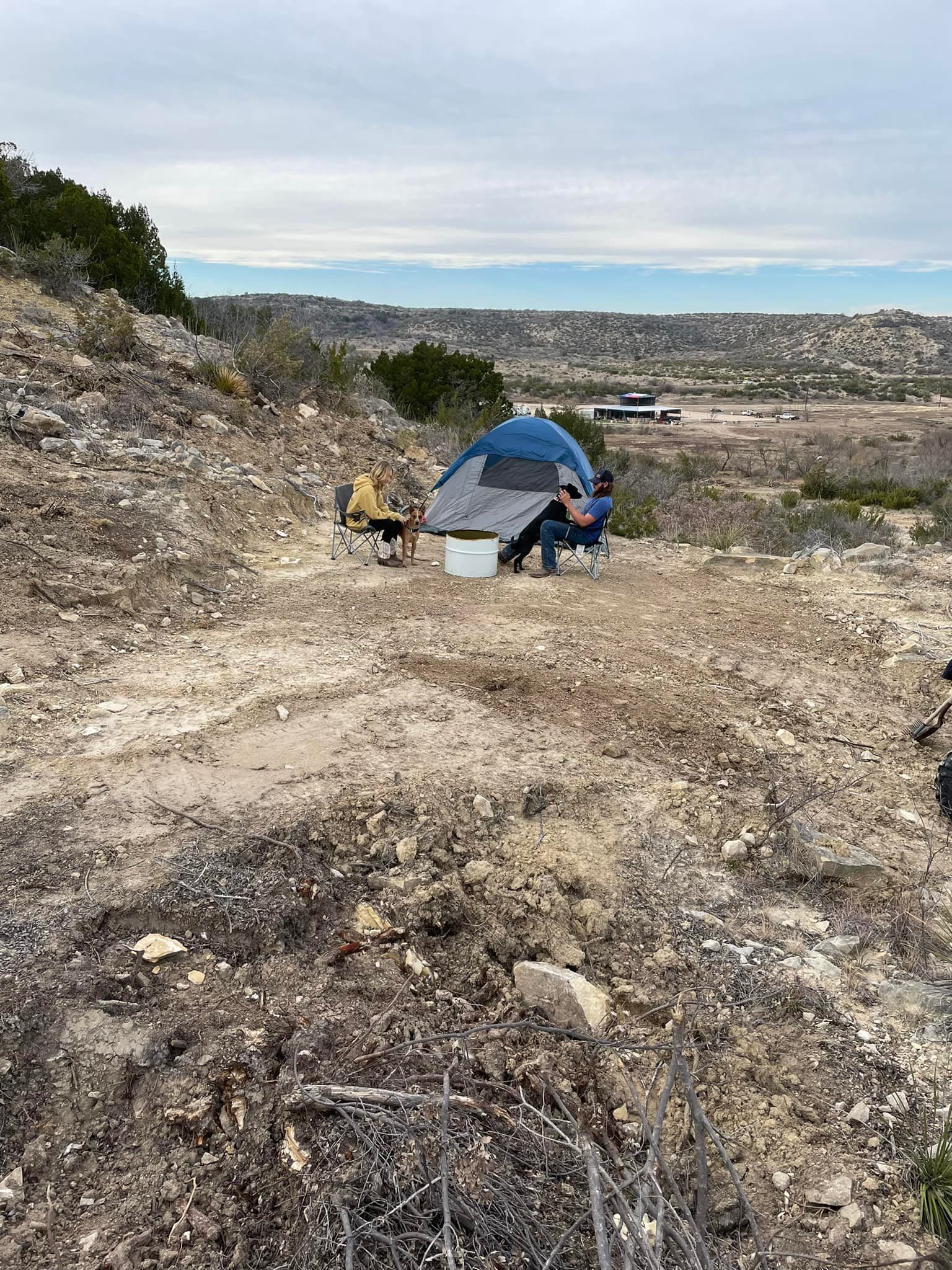 Two people camping beside a blue tent on a rocky hillside overlooking a valley