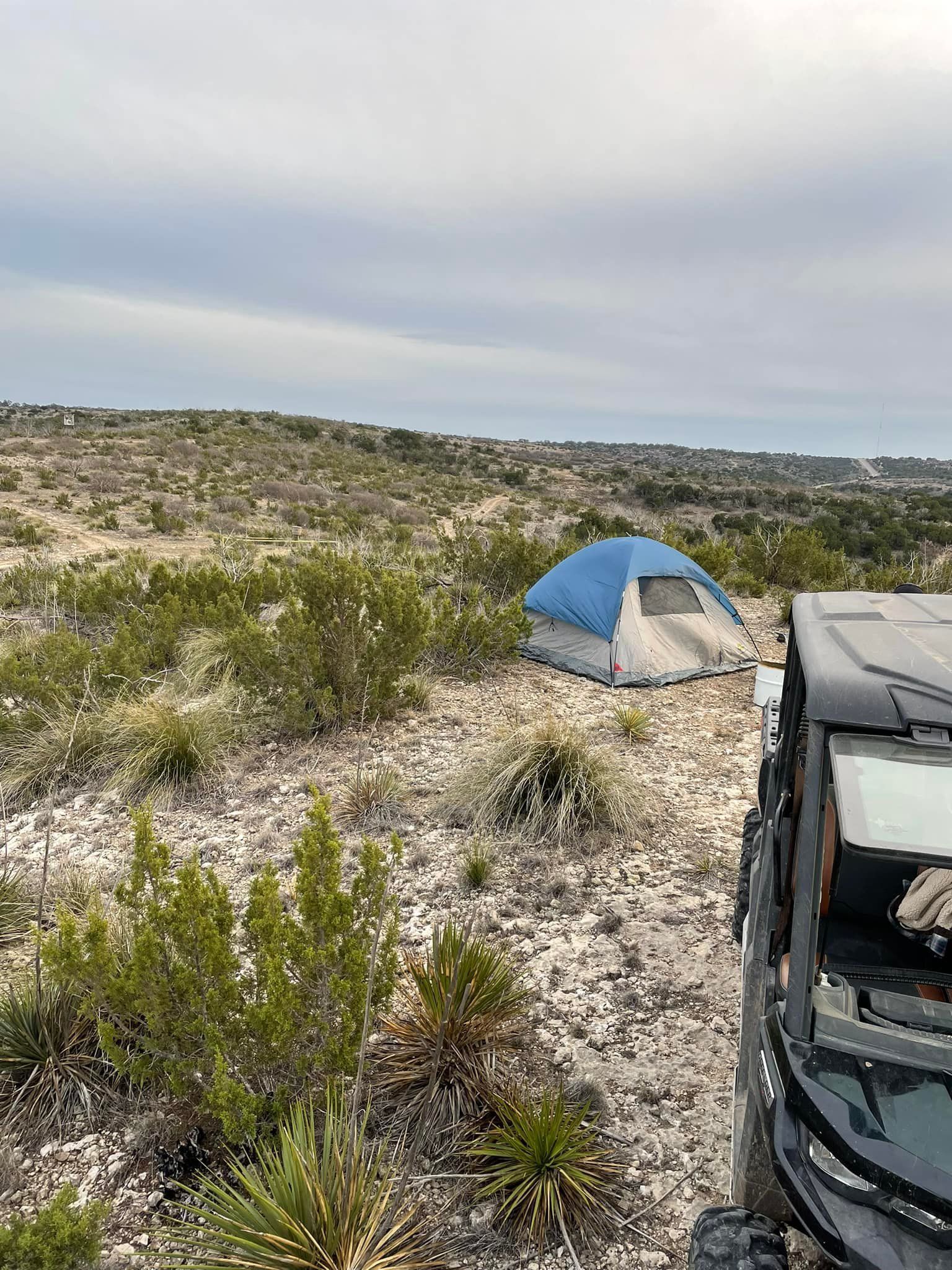 Blue tent on a rocky desert campsite beside a black vehicle under a cloudy sky.