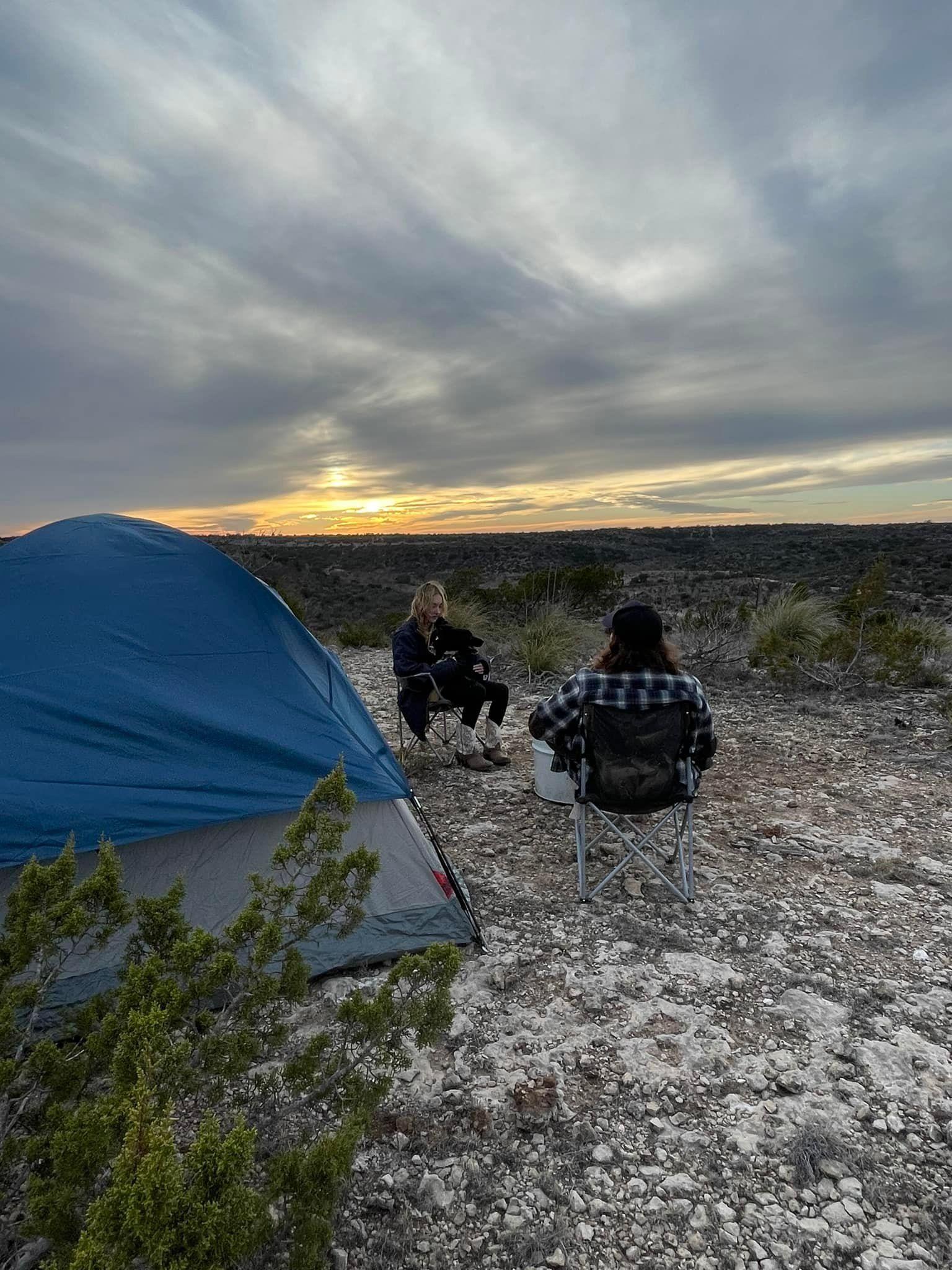 Two campers near a blue tent on a rocky beach at sunset under cloudy skies