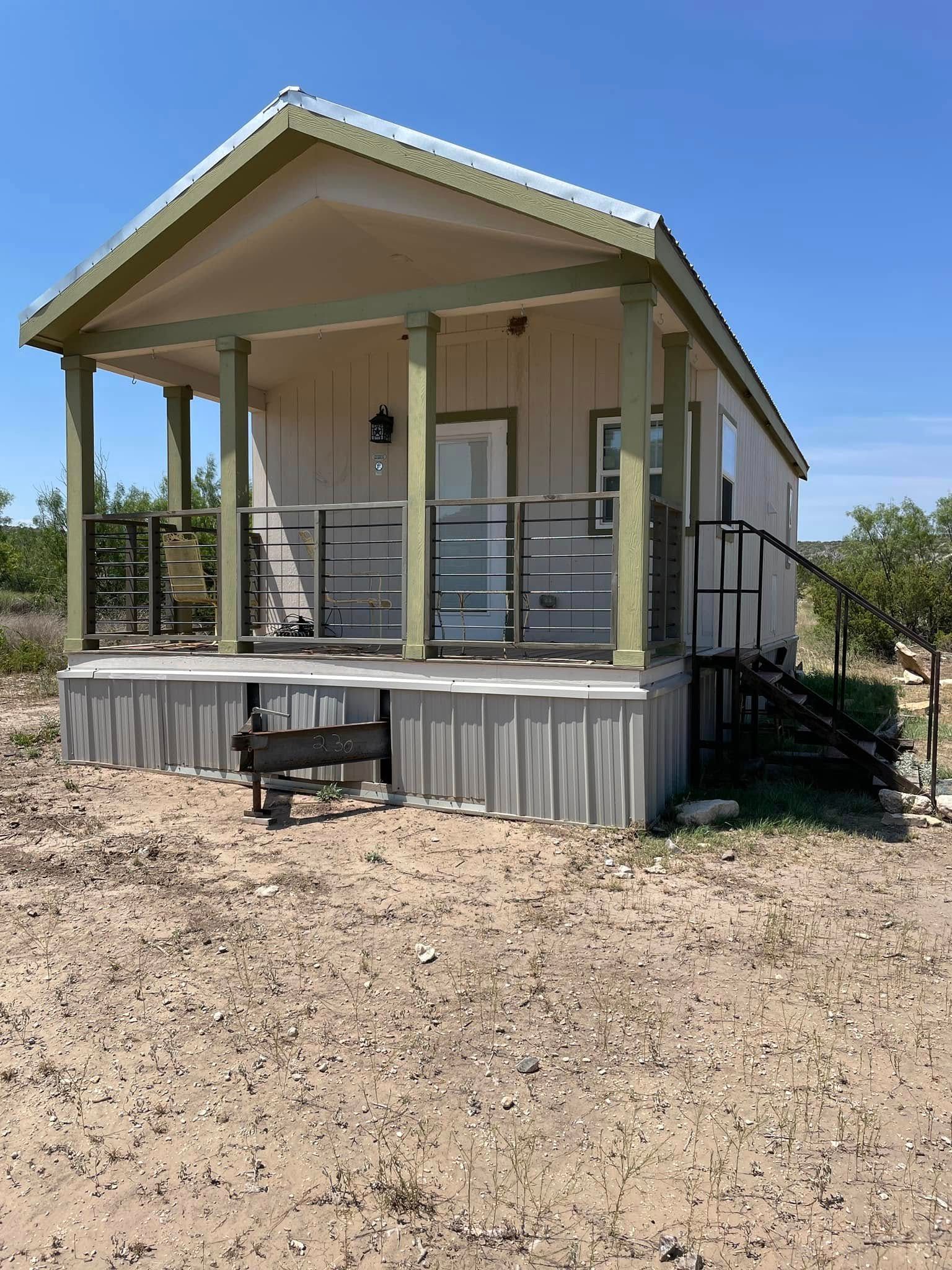 Small beige cabin with porch and ramp, set on a gravel lot under a clear blue sky