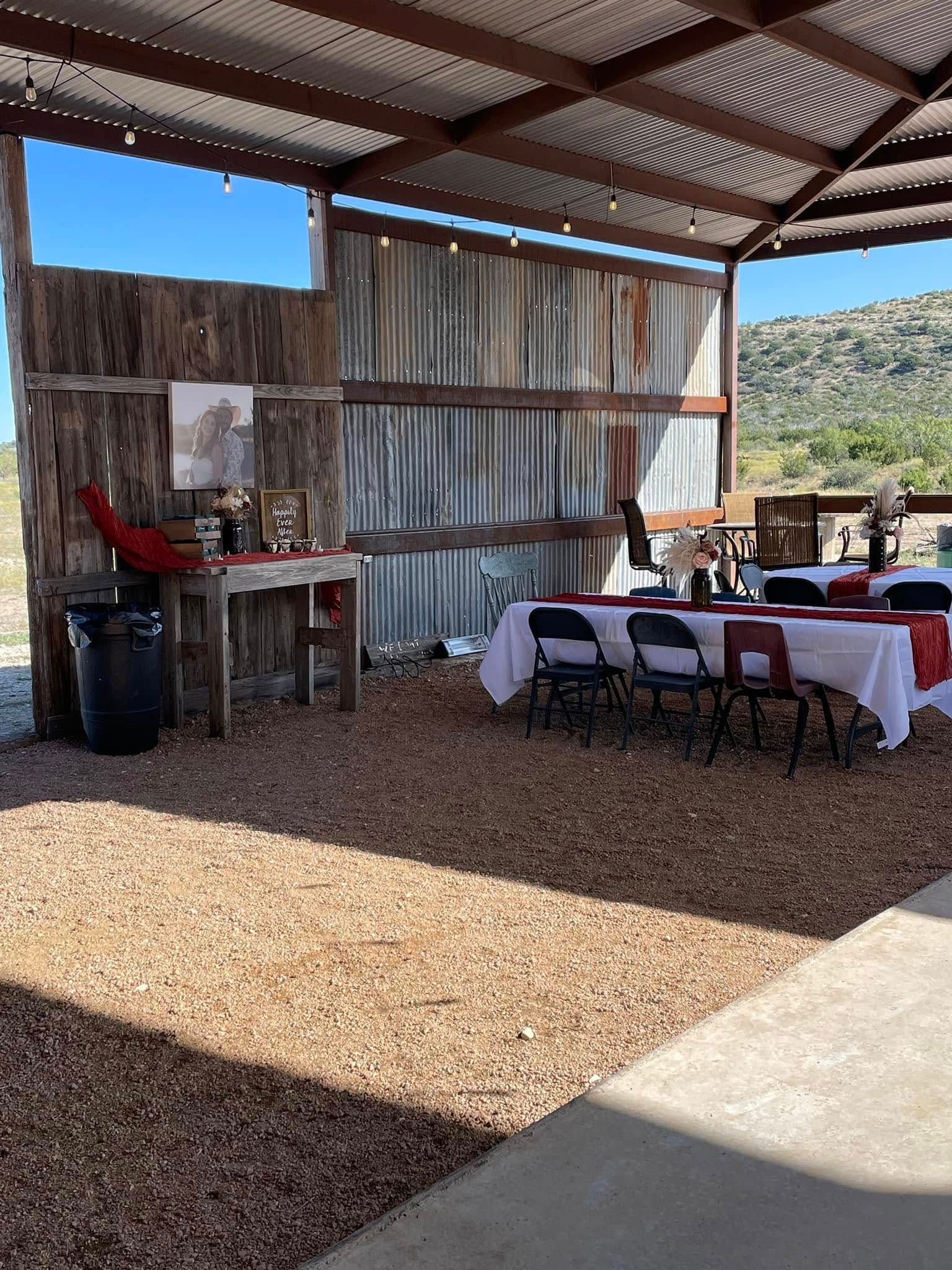 Outdoor rustic shelter with gravel floor, tables, chairs, and a red tablecloth in a bright open-air setting