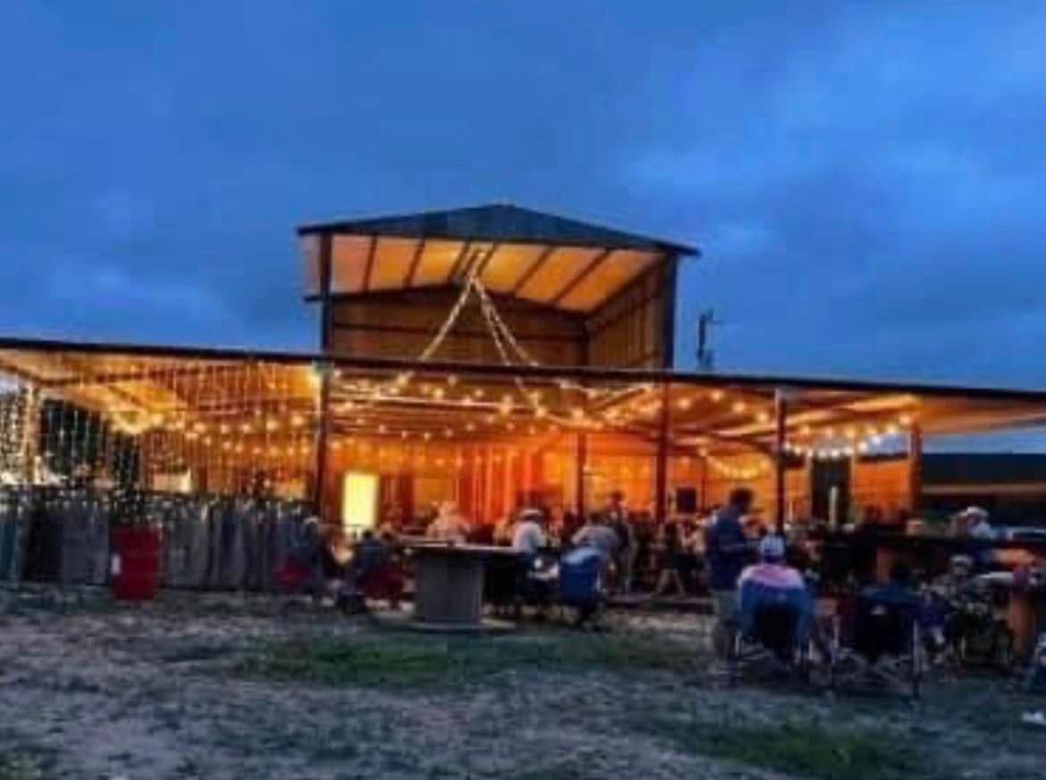 Outdoor evening gathering at a warmly lit rustic pavilion with seated guests under a blue dusk sky