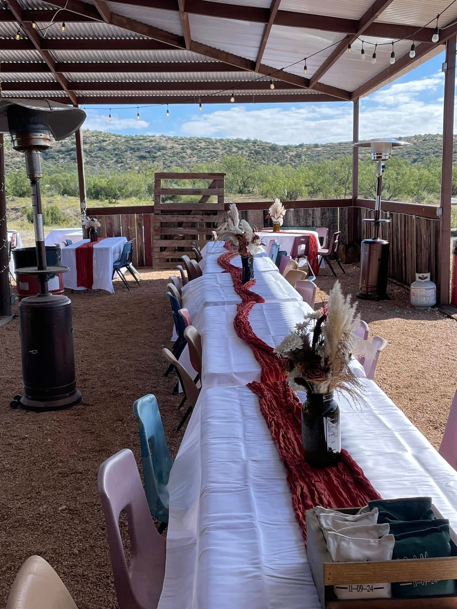 Outdoor banquet table under a pavilion, set with white tablecloths, red runner, and floral centerpieces.