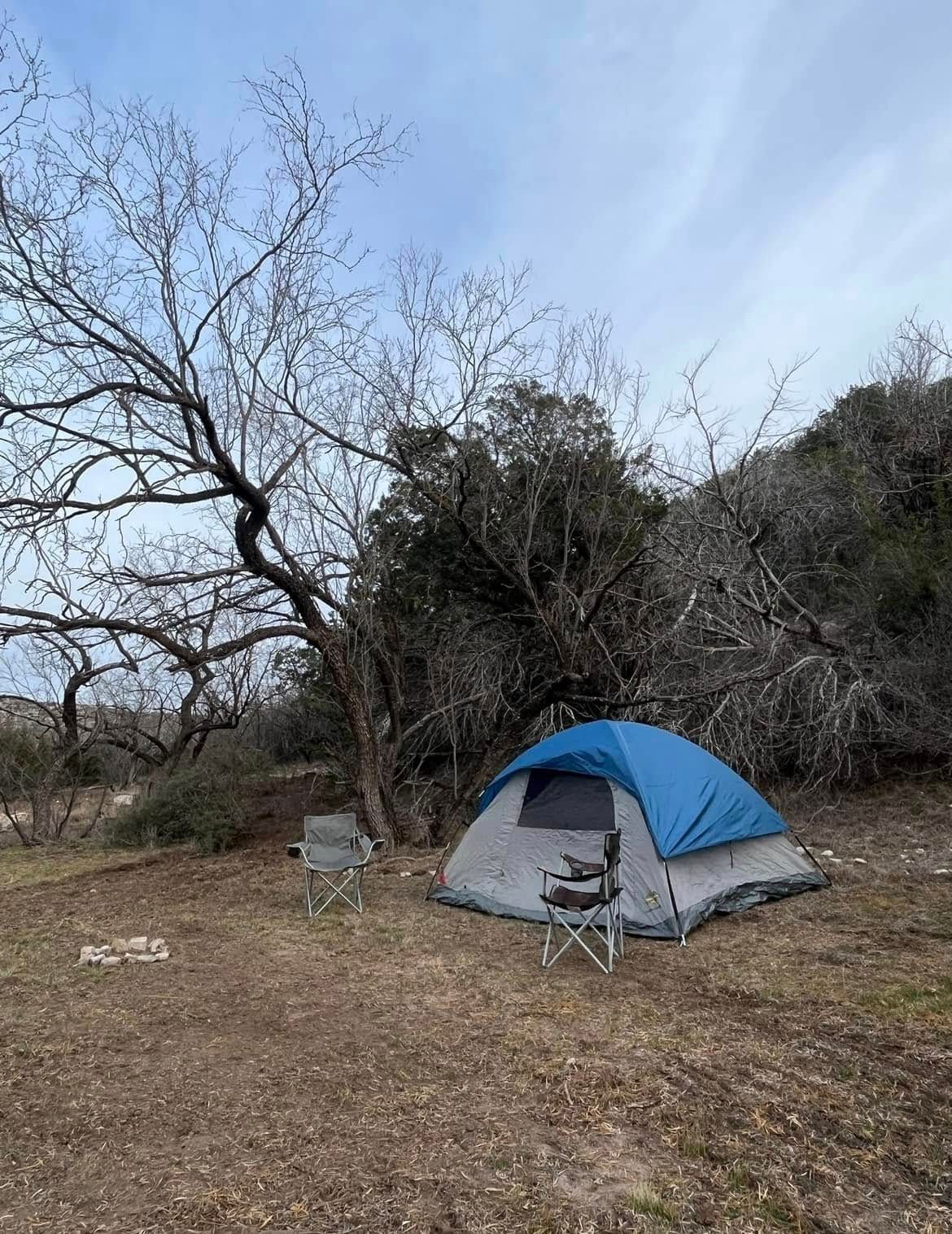 Blue-gray tent and camping chair in a sparse wooded area under a cloudy sky.