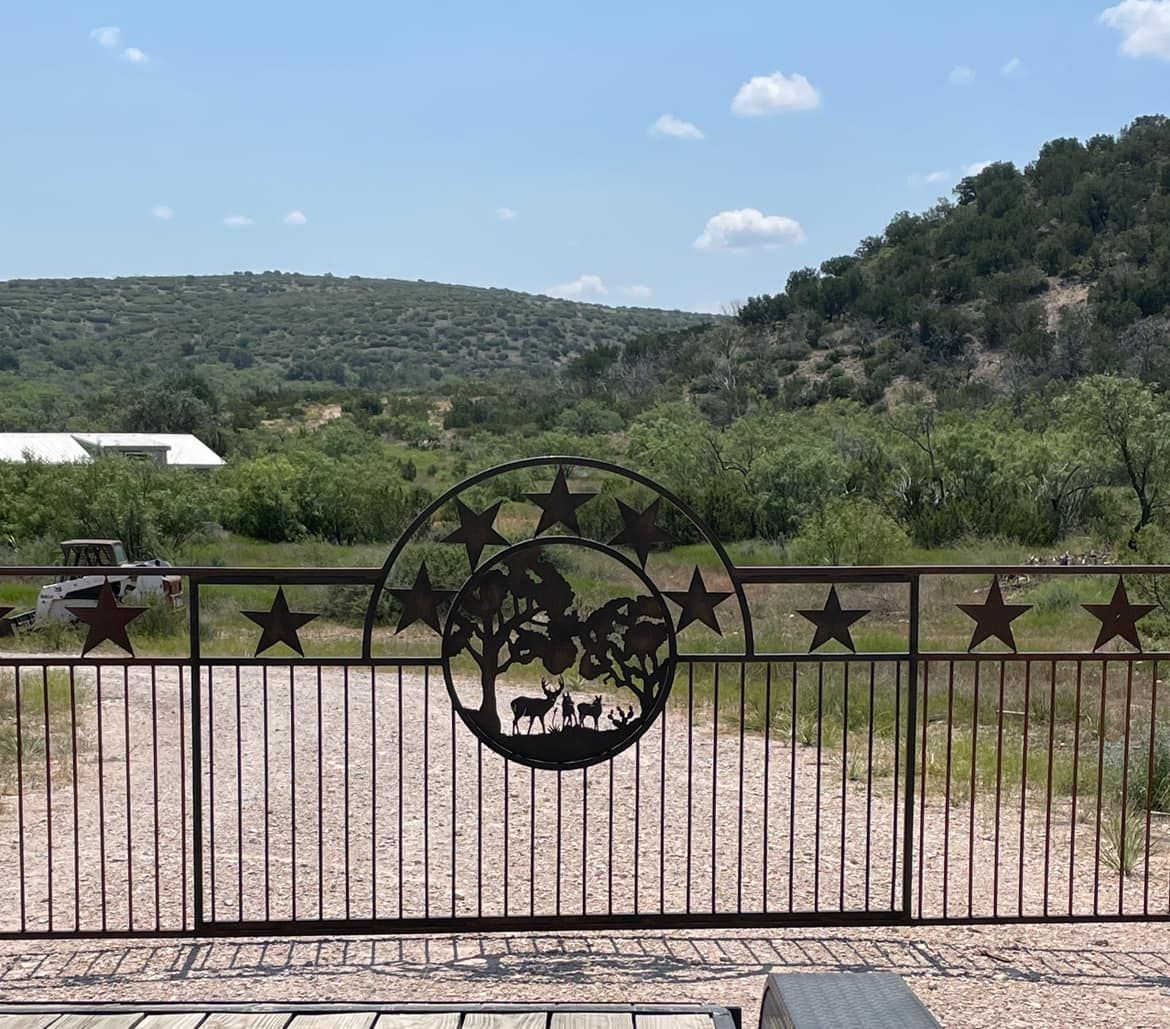 Black metal gate with stars and a circular emblem before a dirt road and green hillside under a blue sky