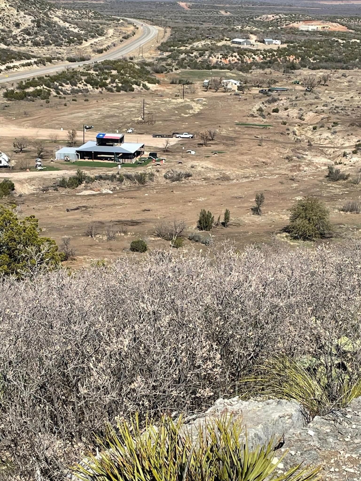 Rural hillside with a small building, dry open fields, and a road in the distance under a cloudy sky