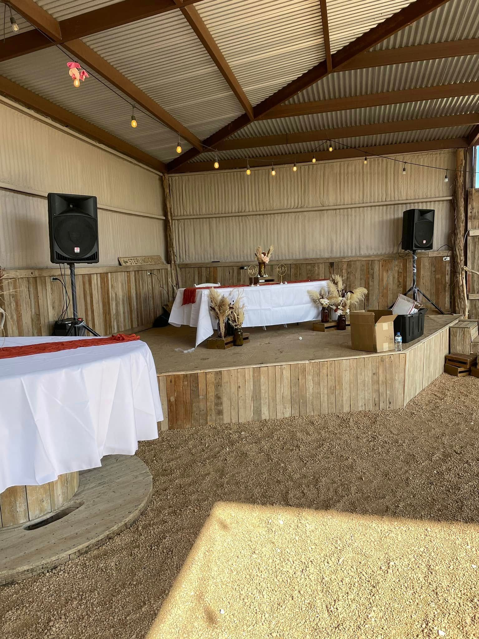 Rustic indoor event hall with a decorated stage, tables, and speakers under a wooden roof