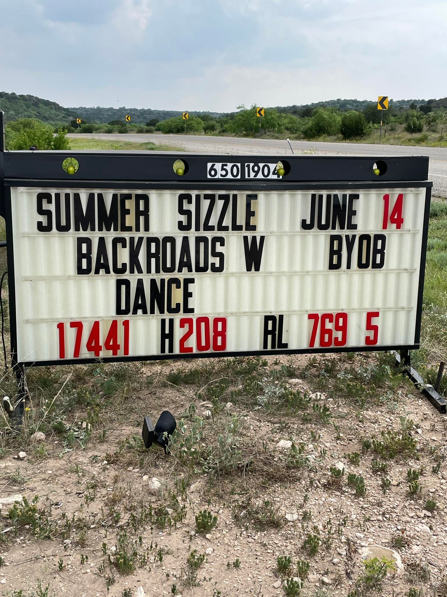 Outdoor marquee sign reading “Summer Sizzle,” “Backroads,” “Dance,” “Junk,” and event details in a grassy field.