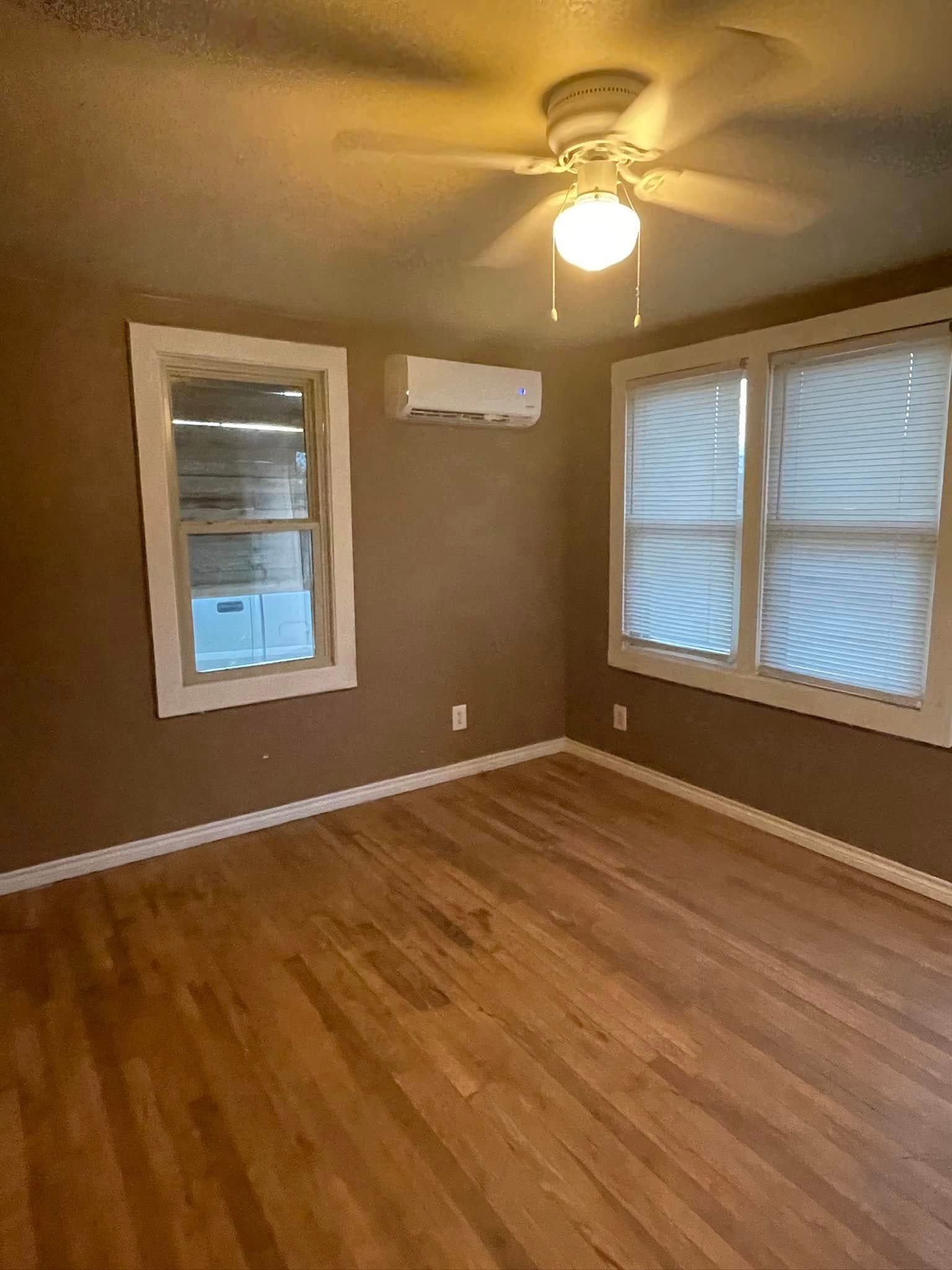 Empty room with wood floors, beige walls, windows, ceiling fan, and wall-mounted air conditioner.