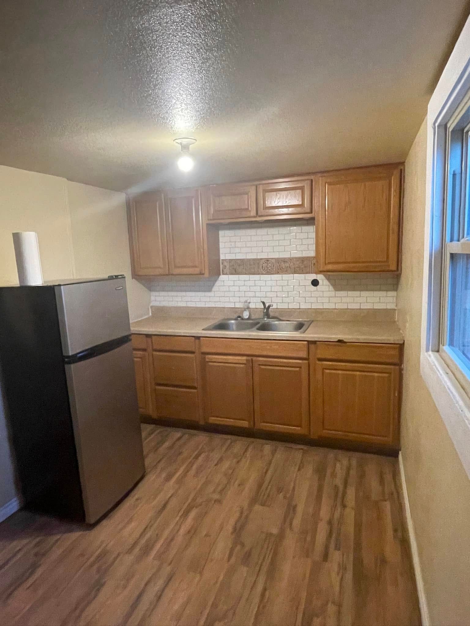 Small kitchen with wood cabinets, white tile backsplash, black refrigerator, and wood-look floor.