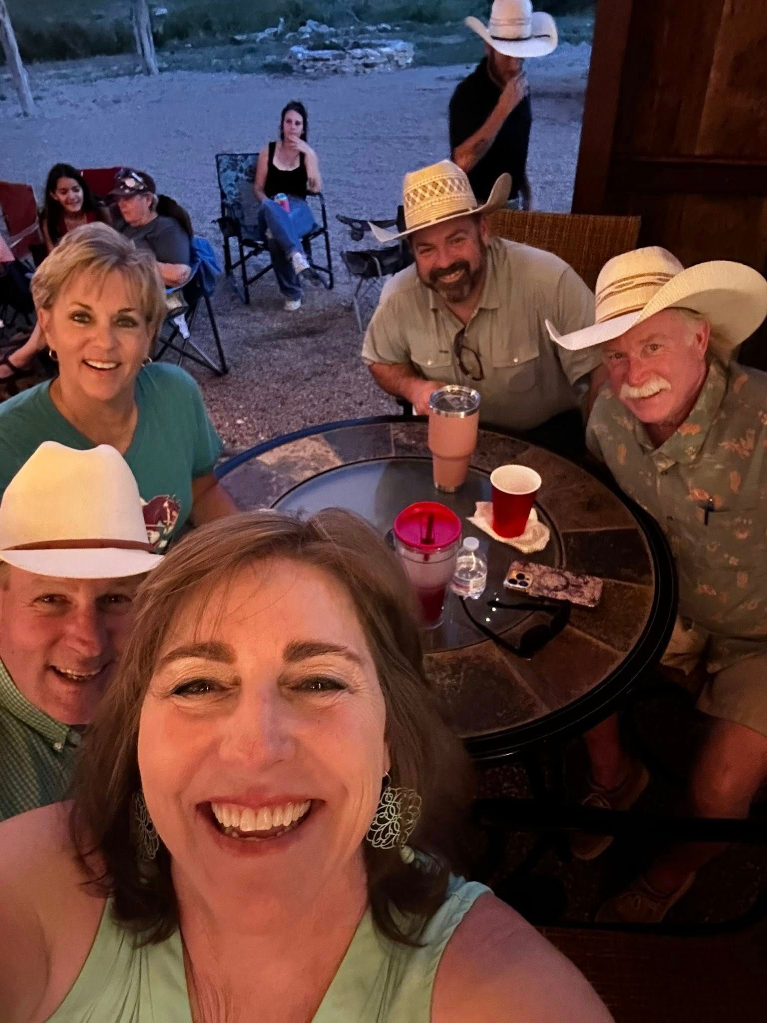 Smiling group seated around a table outdoors at dusk, with cowboy hats and drinks on the table.