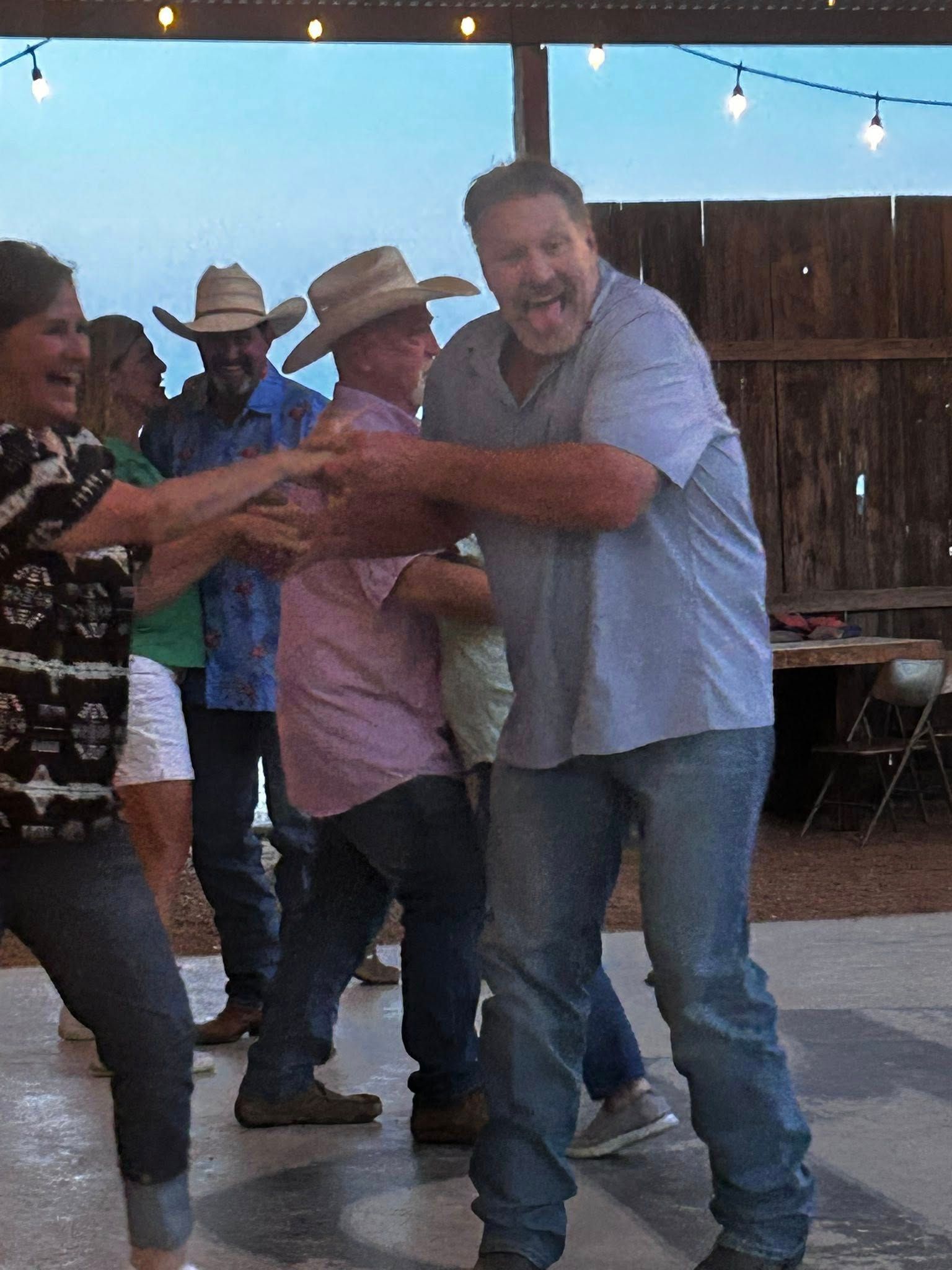 People dancing at an outdoor party under string lights, with a man in a light blue shirt in front.