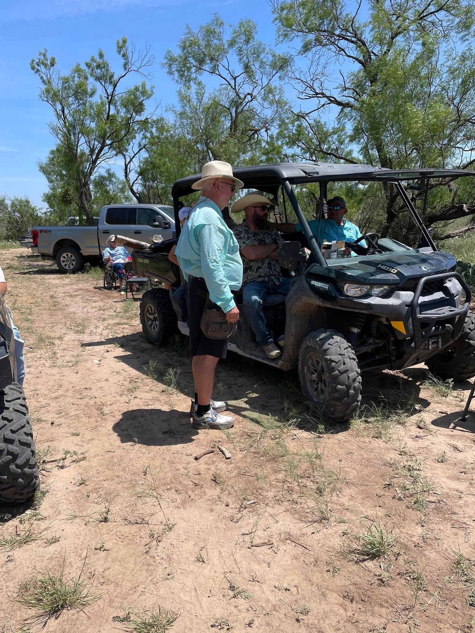 Two people by a black off-road vehicle on a dirt trail, with a truck and trees in the background.