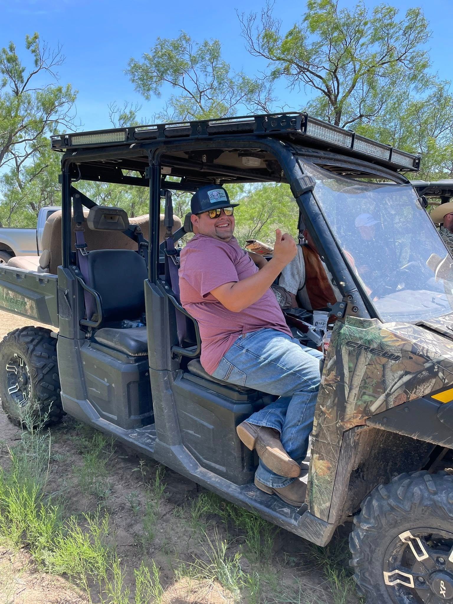 Man driving a muddy off-road utility vehicle on a grassy trail under a clear blue sky.