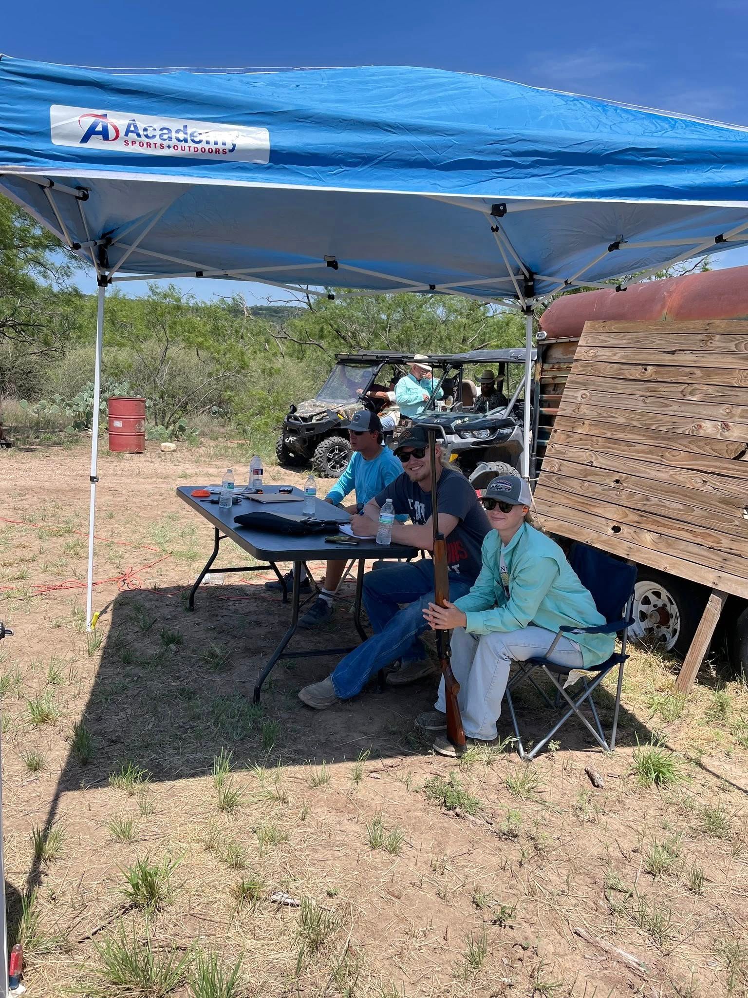 Two people sitting under a blue canopy at an outdoor table, with a stack of hay bales nearby.