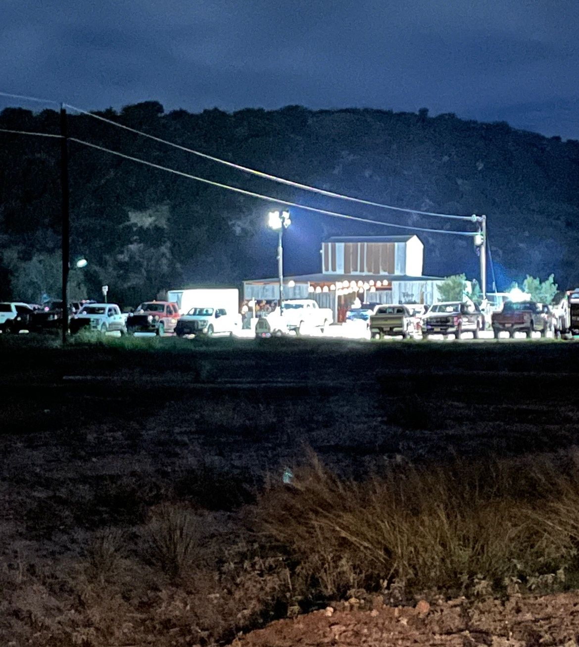Nighttime roadside scene with lit storefronts, parked cars, and a wooded hill in the background.