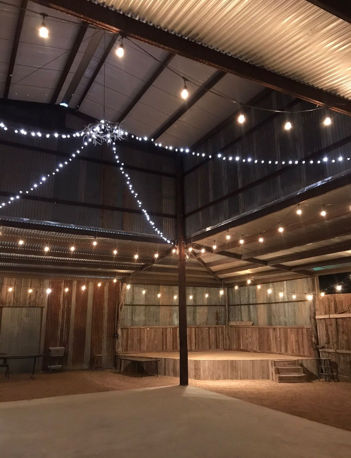 String lights hang under a metal-roofed pavilion with a central pole and empty concrete floor.