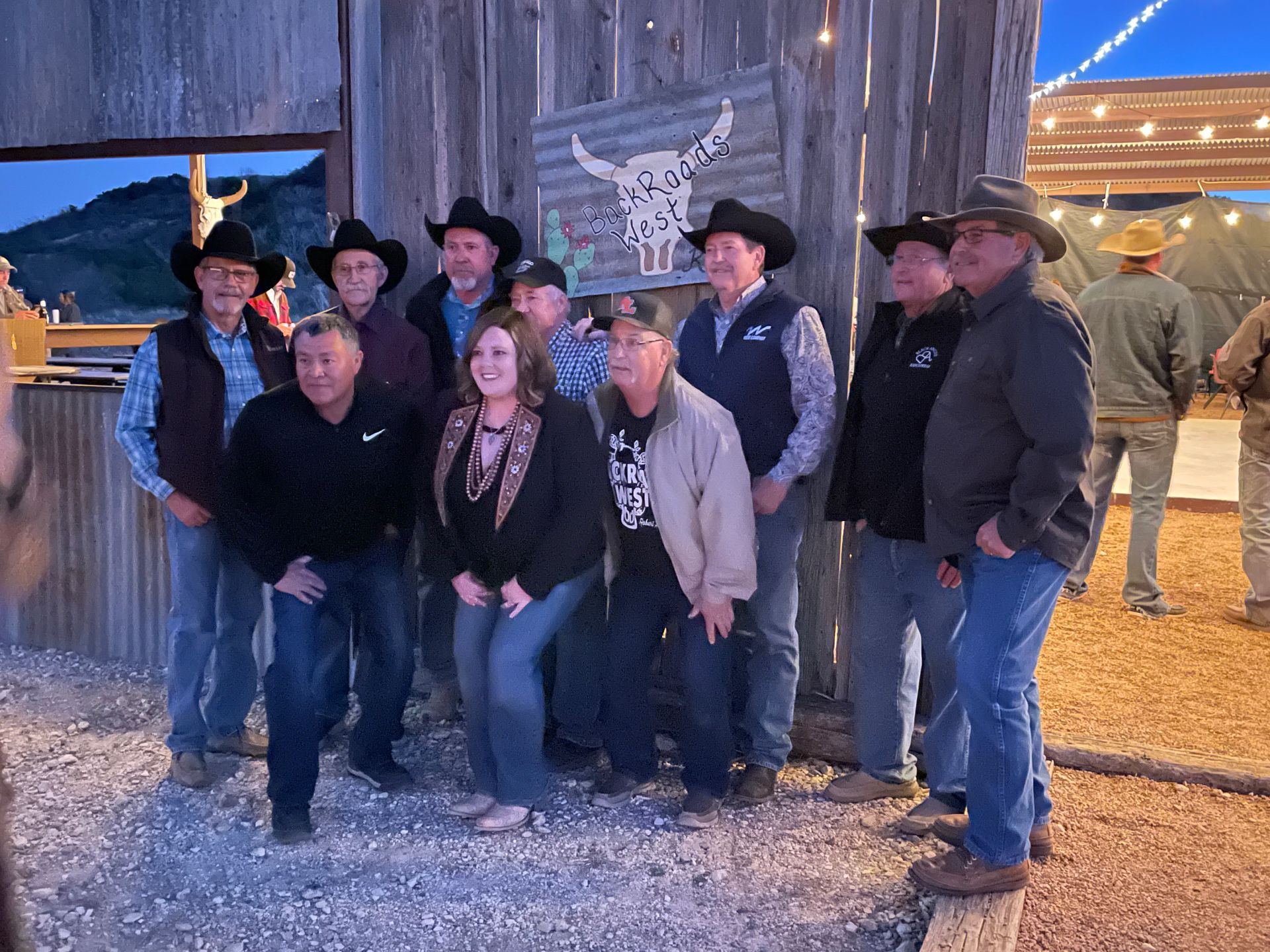 Group posing outdoors at night in cowboy hats beside a lit barn and string lights.