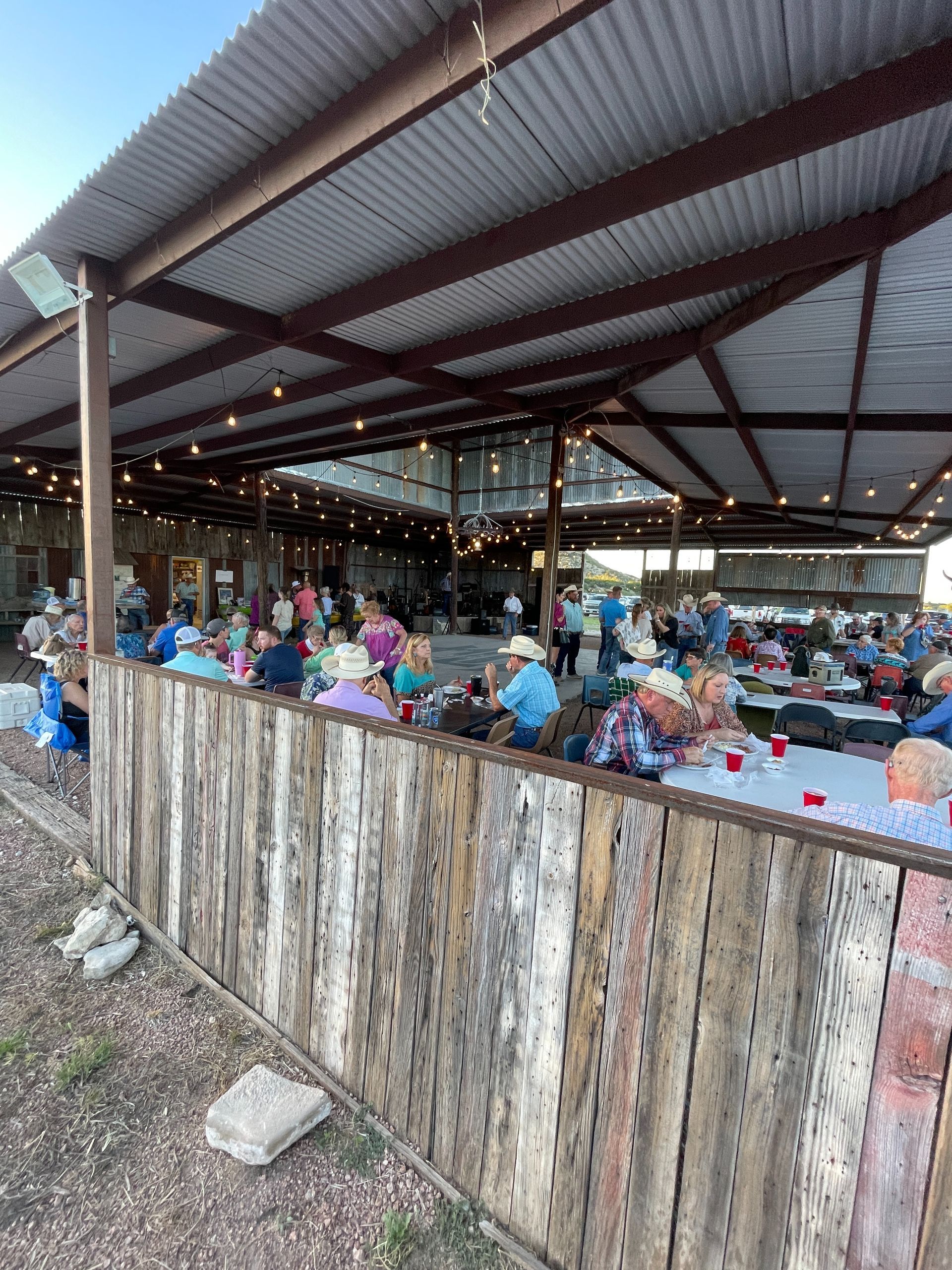Open-air dining area with many people seated under a metal roof behind a rustic wooden fence