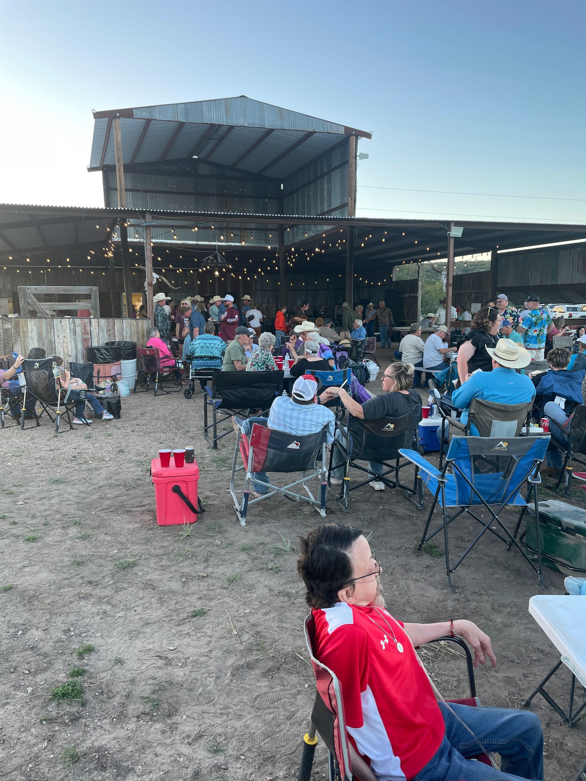Outdoor concert crowd seated in folding chairs at dusk, facing a large stage with a covered roof.