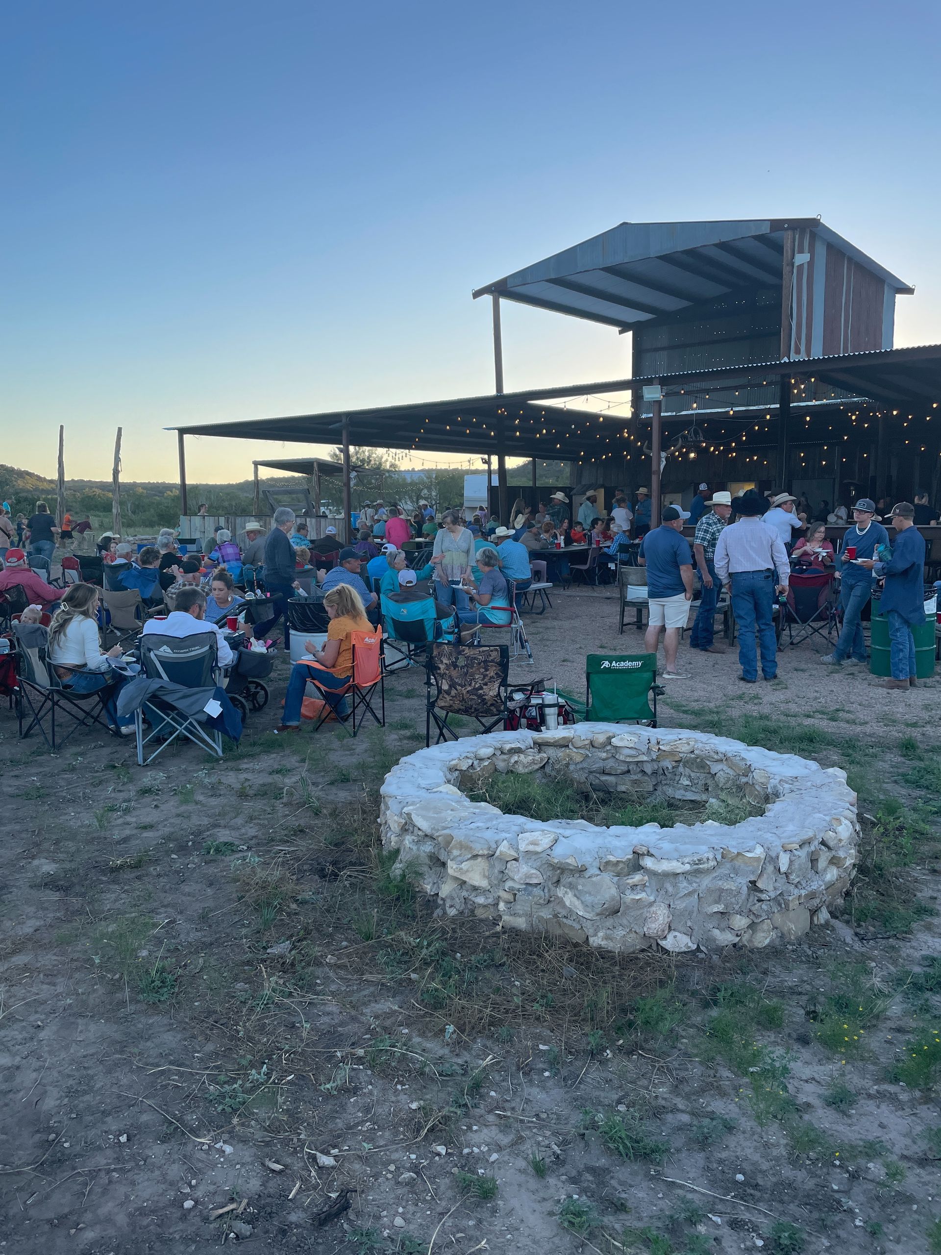 Crowd seated outdoors at a pavilion at dusk, with a white stone planter in the foreground.