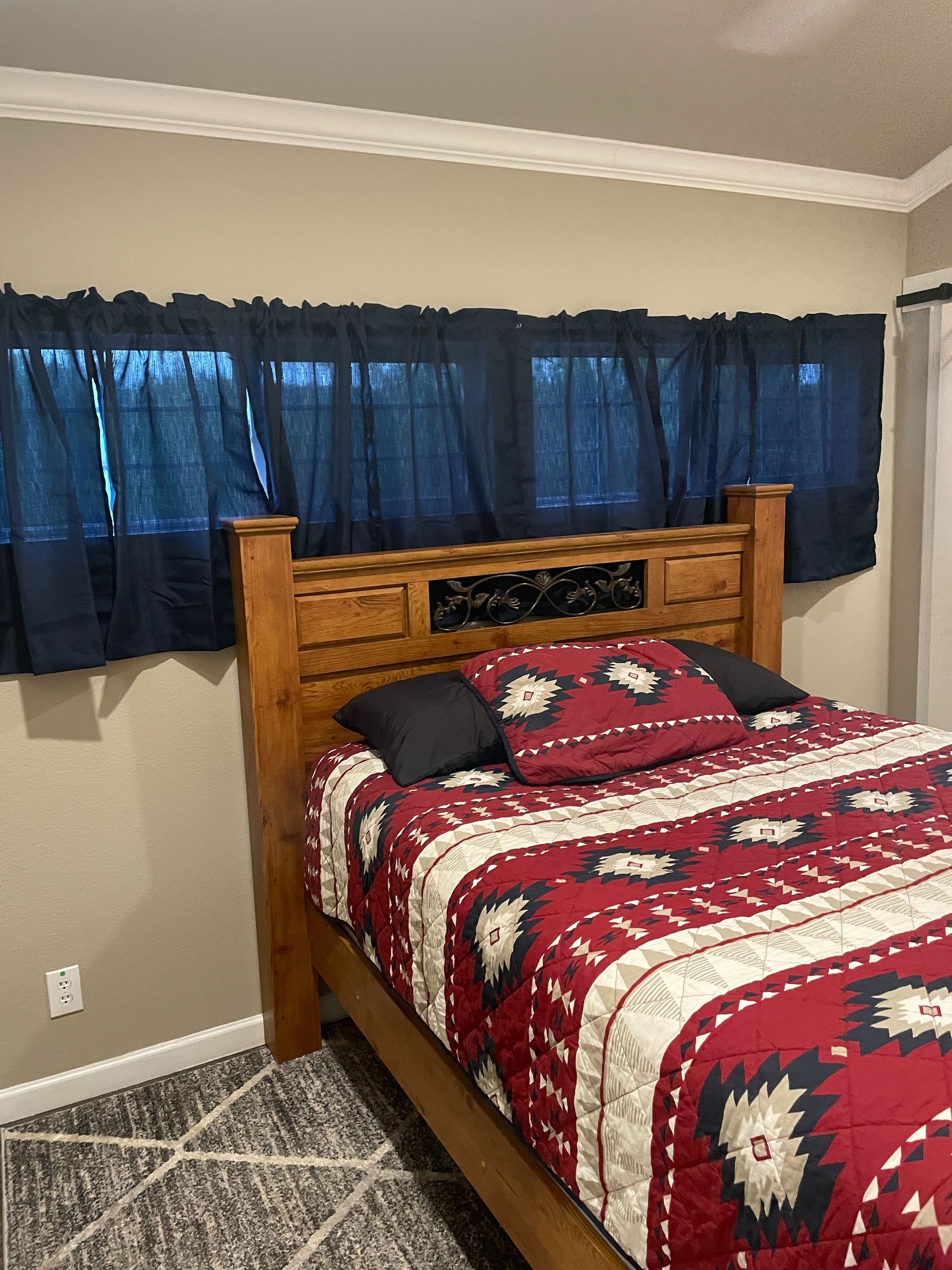 Bedroom with wooden bed, red patterned quilt, blue curtains, and gray carpet.