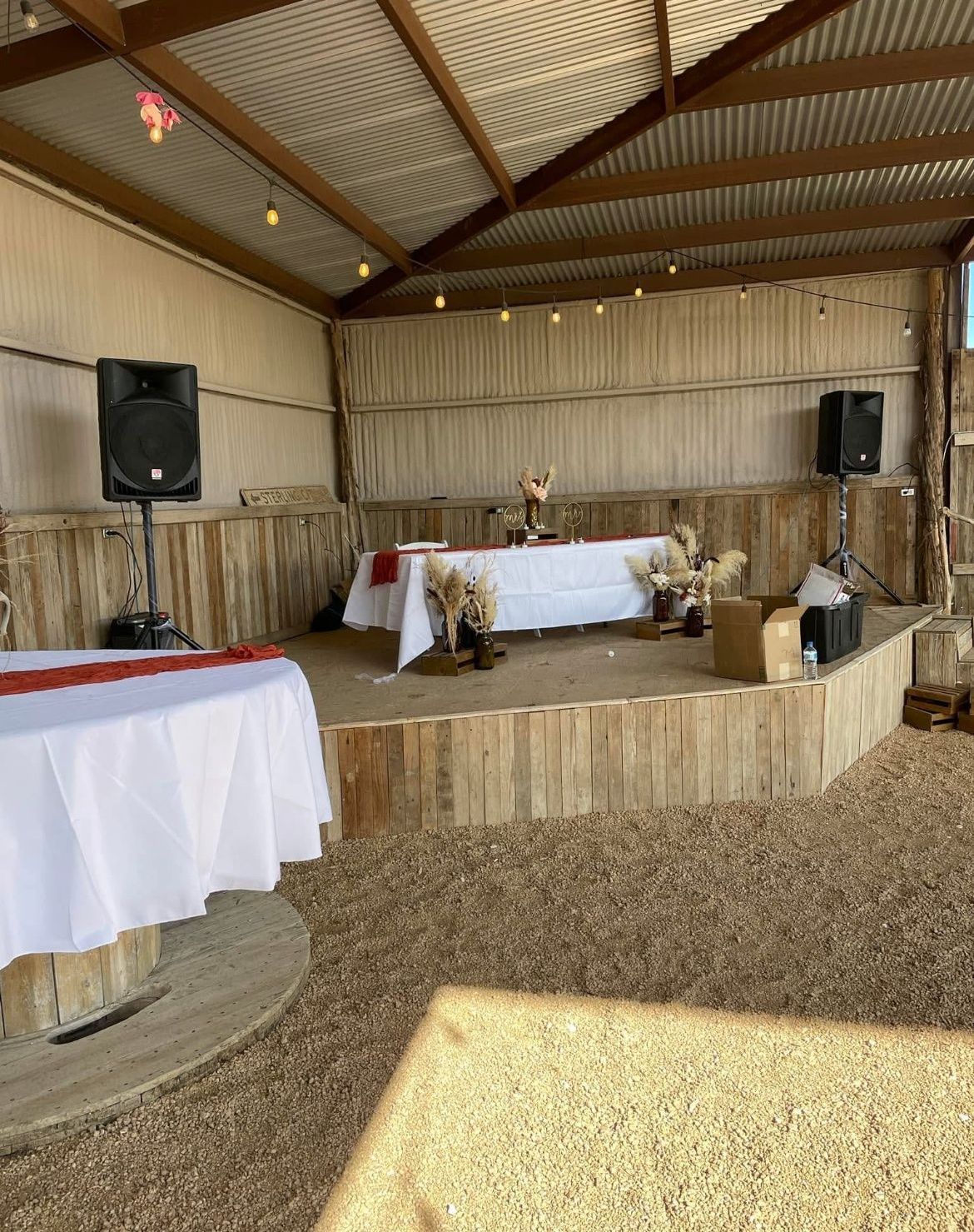Rustic indoor event stage with white-draped tables, speakers, and string lights under a wooden roof