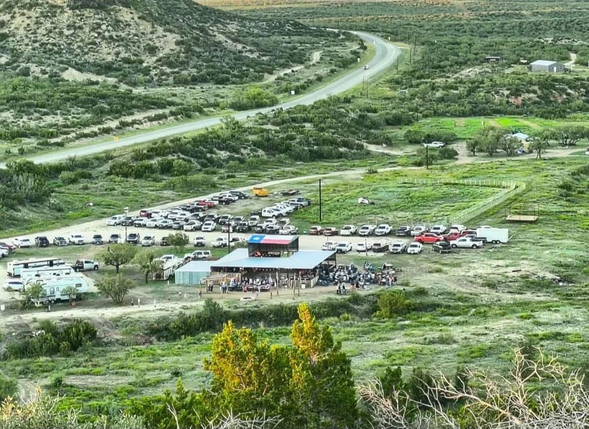 Crowded roadside rest stop in a green valley with parked cars and a winding road in the background