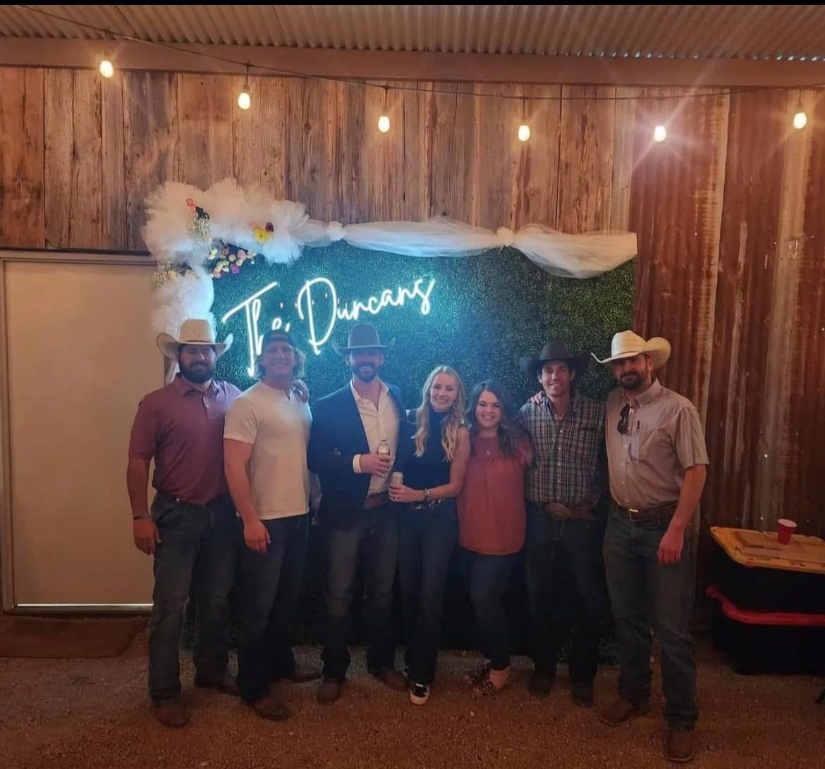 Group of six posing at a rustic indoor event with a neon sign and white draped decor behind them.