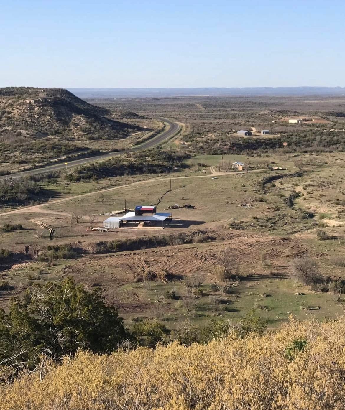 Rural desert landscape with a winding dirt road, low hills, and a small building under a clear blue sky