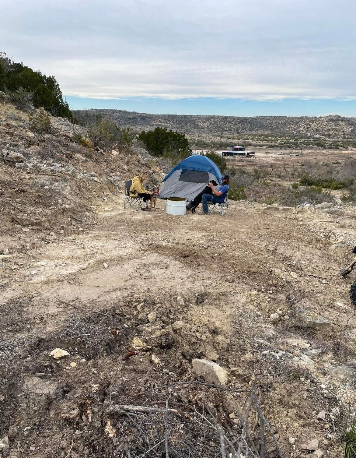 Two people camping beside a blue tent in a rocky desert landscape under a cloudy sky