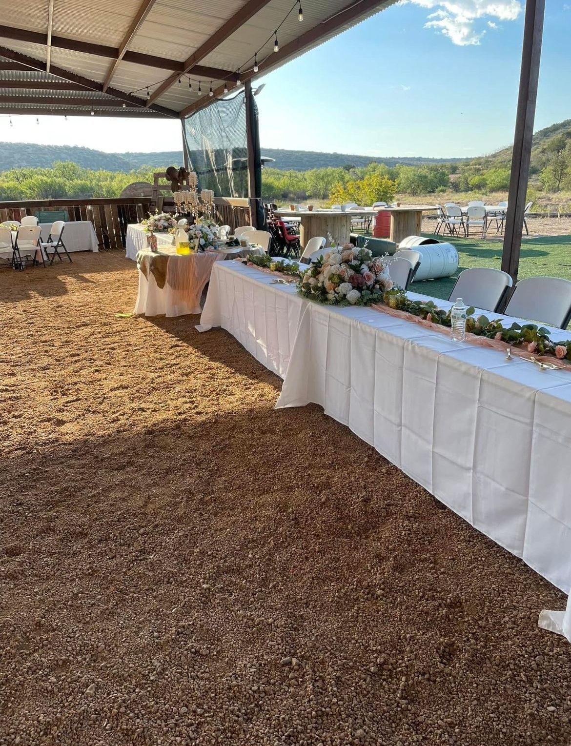 Outdoor buffet table with white tablecloths under a canopy, set against a sunny landscape.