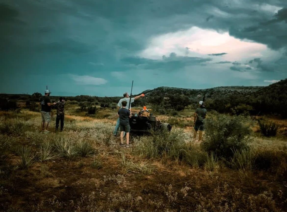 People in a dry field under a stormy sky, gathered around a vehicle or equipment.