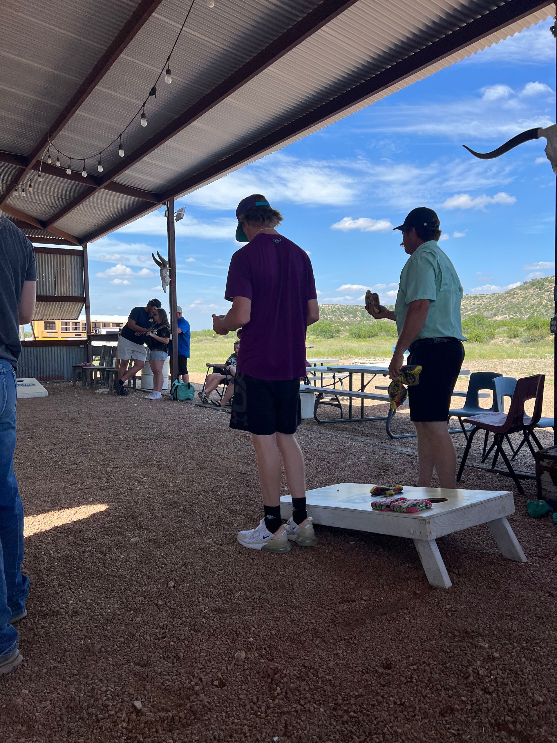 People standing under a shaded pavilion near benches and a cornhole board on a sunny day.