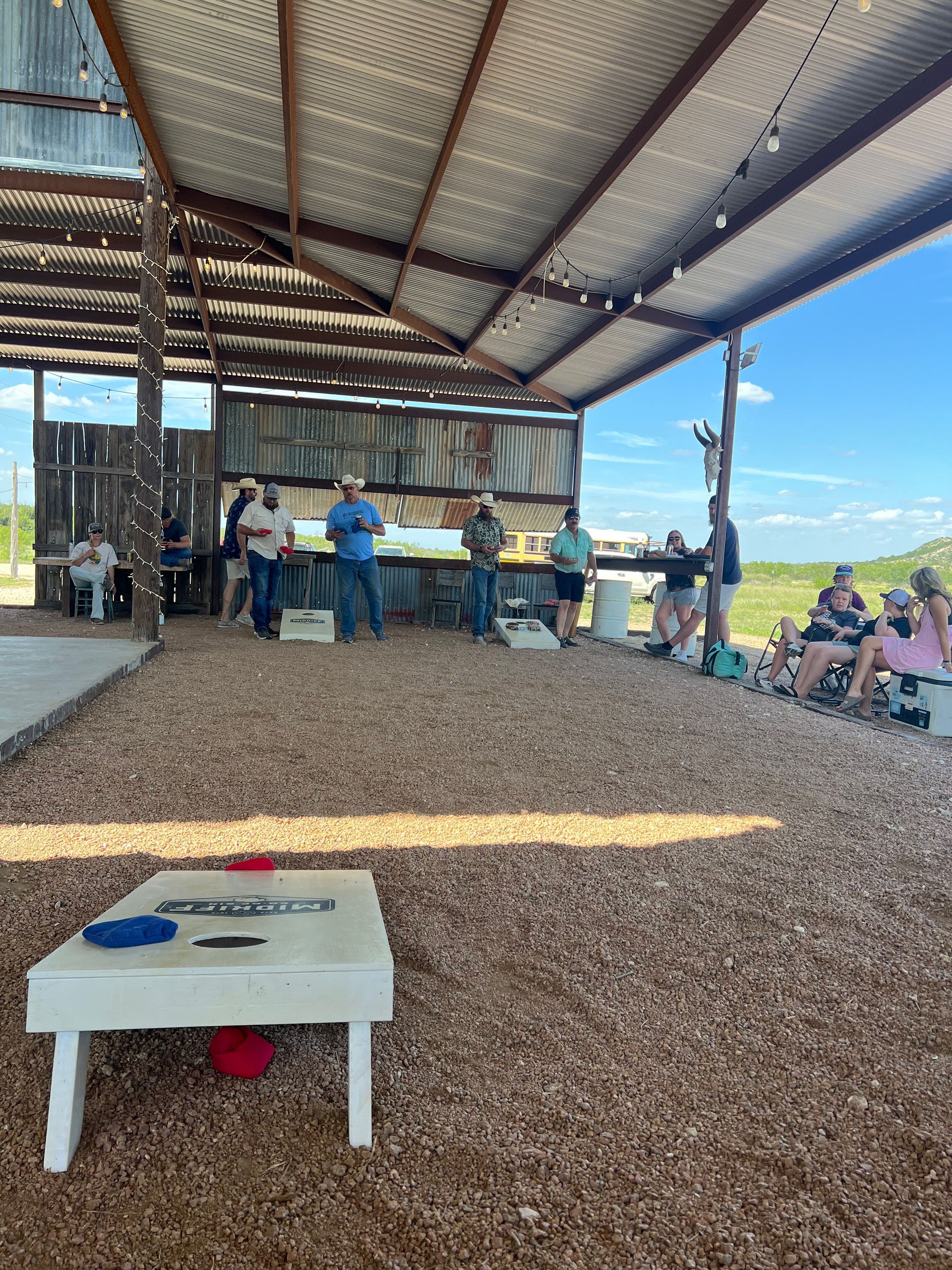 Open-air pavilion with people gathered at a counter, gravel floor, and a cornhole board in the foreground