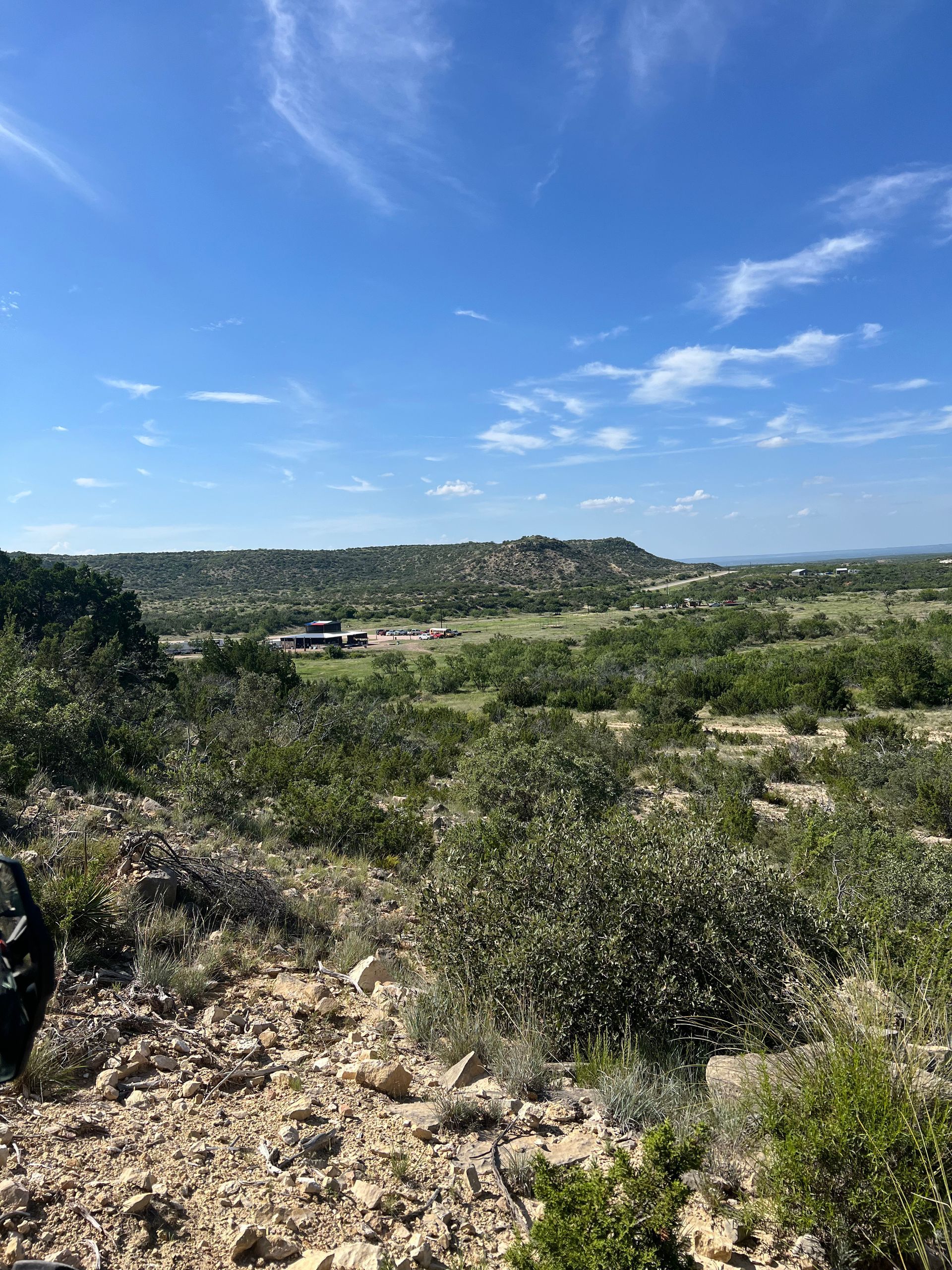 Rocky desert landscape with green shrubs, distant hills, and a bright blue sky with scattered clouds
