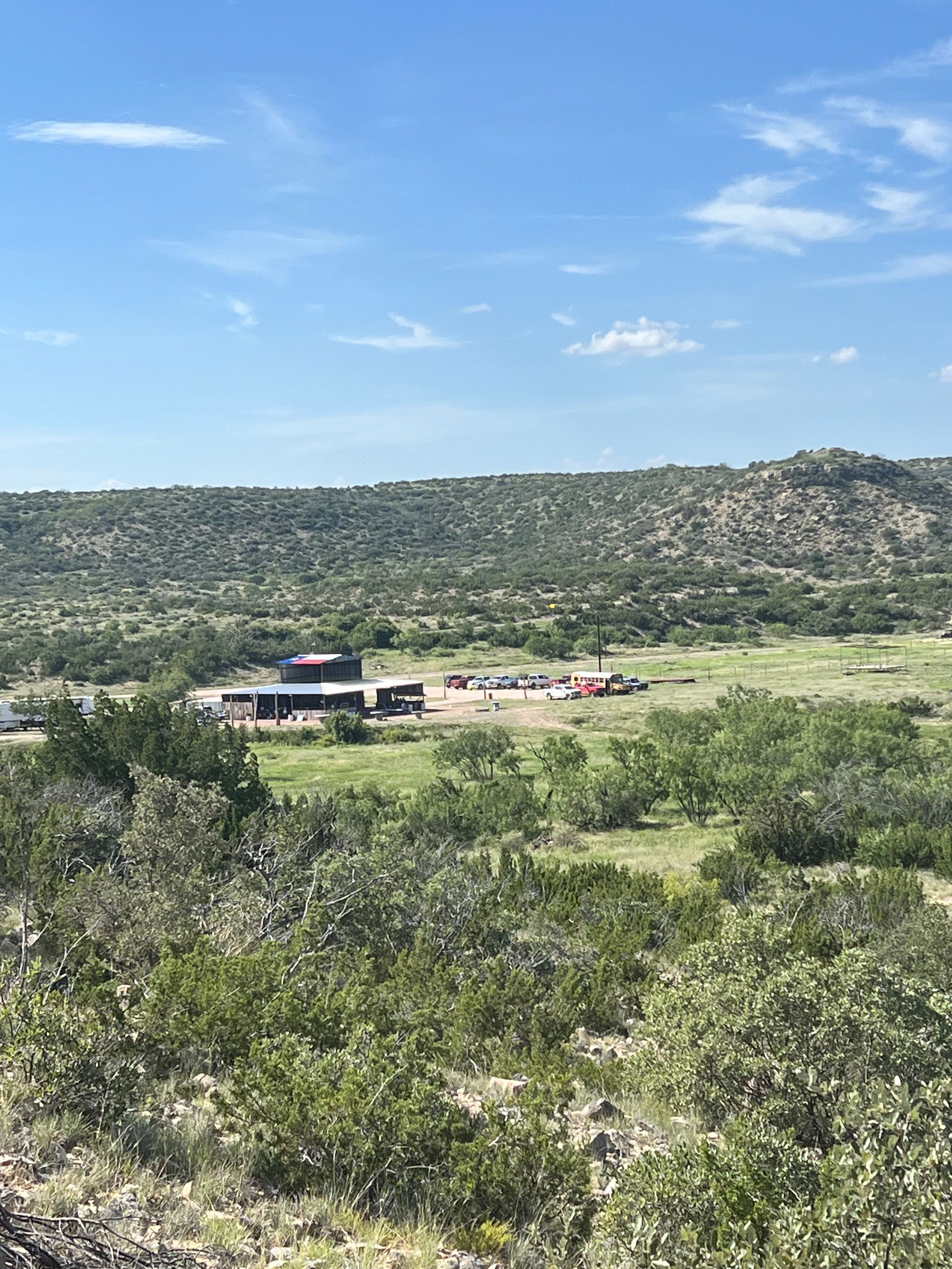 Wide rural landscape with green scrubland, a small building, and distant hills under a blue sky.