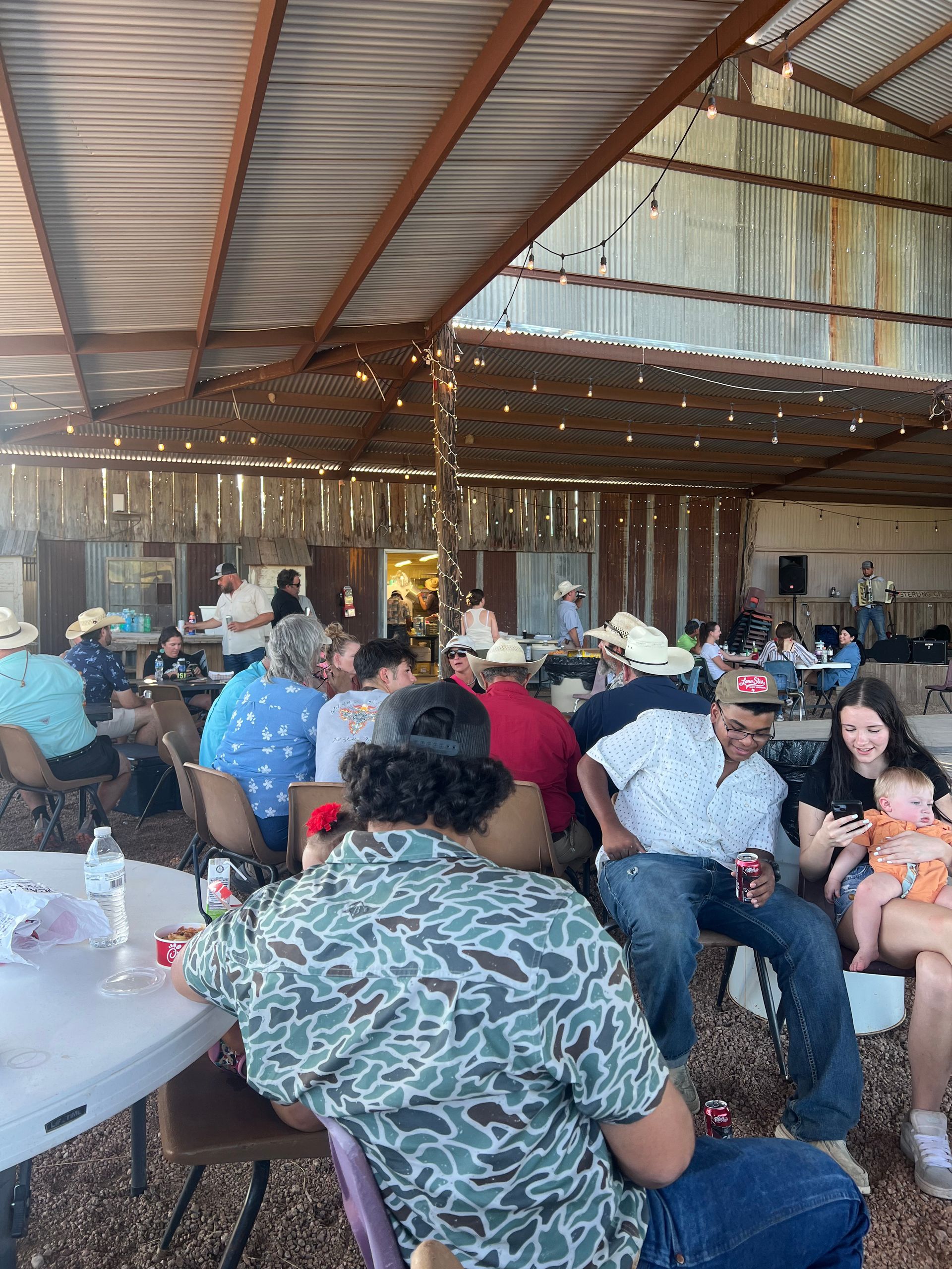 People seated at tables inside a rustic barn-like event hall with string lights and a corrugated metal roof.