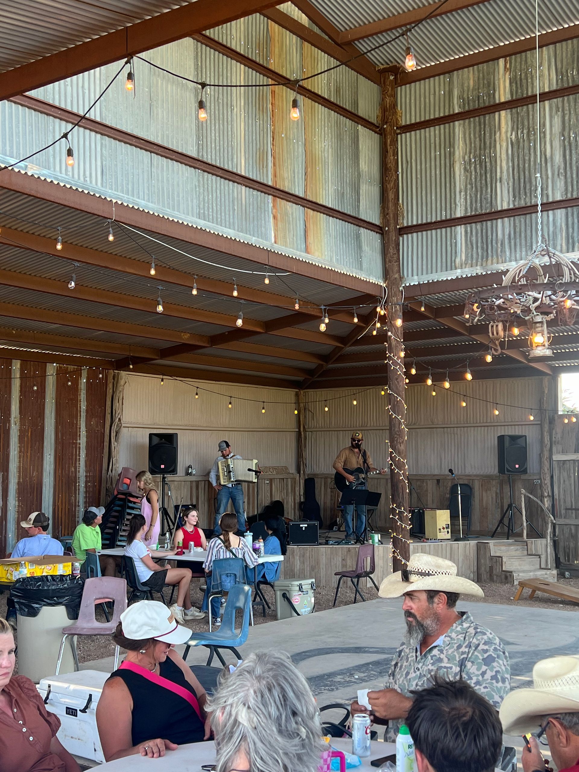 People seated in a rustic barn venue, facing a live band on a small stage with string lights.