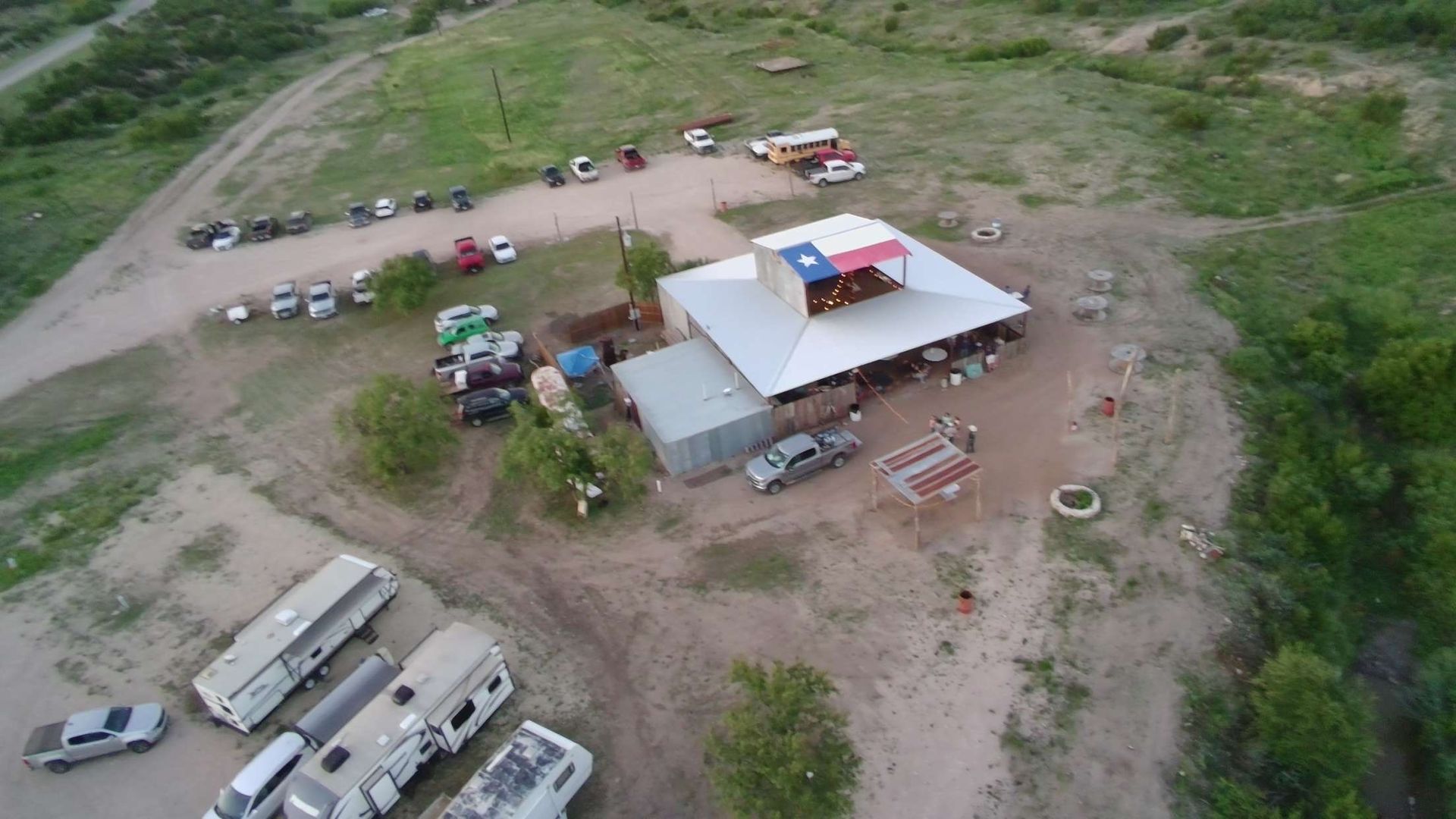 Aerial view of a rural campsite with tents, vehicles, and a central shelter on grassy terrain.