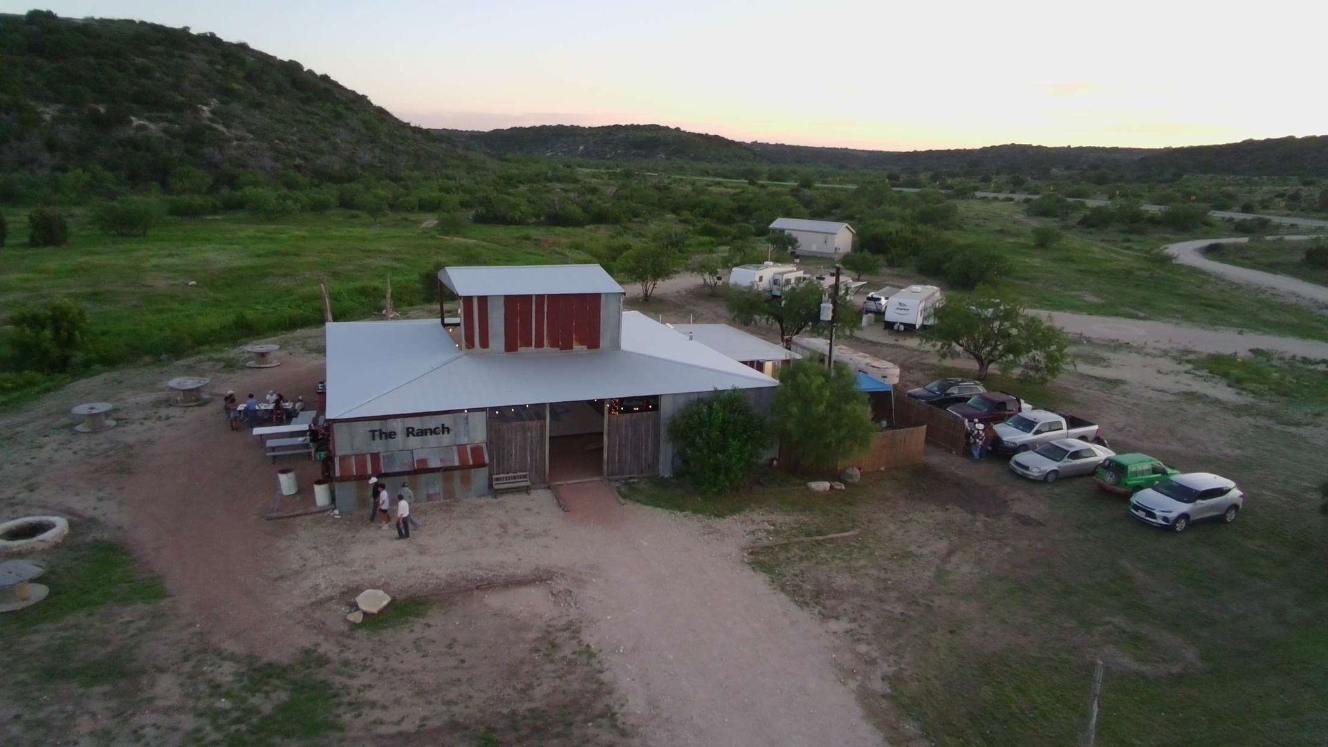 Rural lodge at dusk with parked cars, dirt lot, and green hills in the background