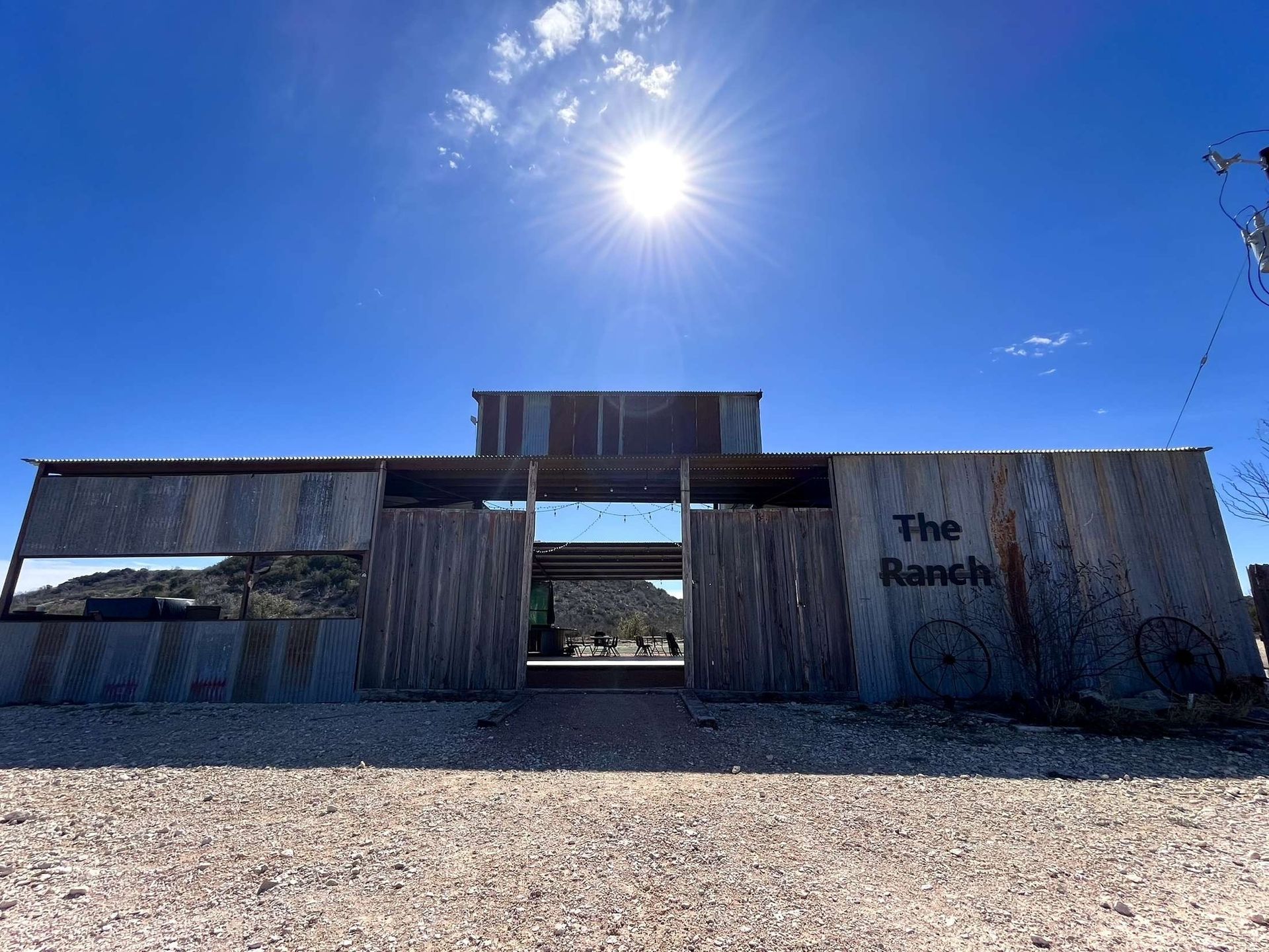 Rustic corrugated-metal ranch gate under a bright sun in a gravel lot, with “The Ranch” painted on the right wall
