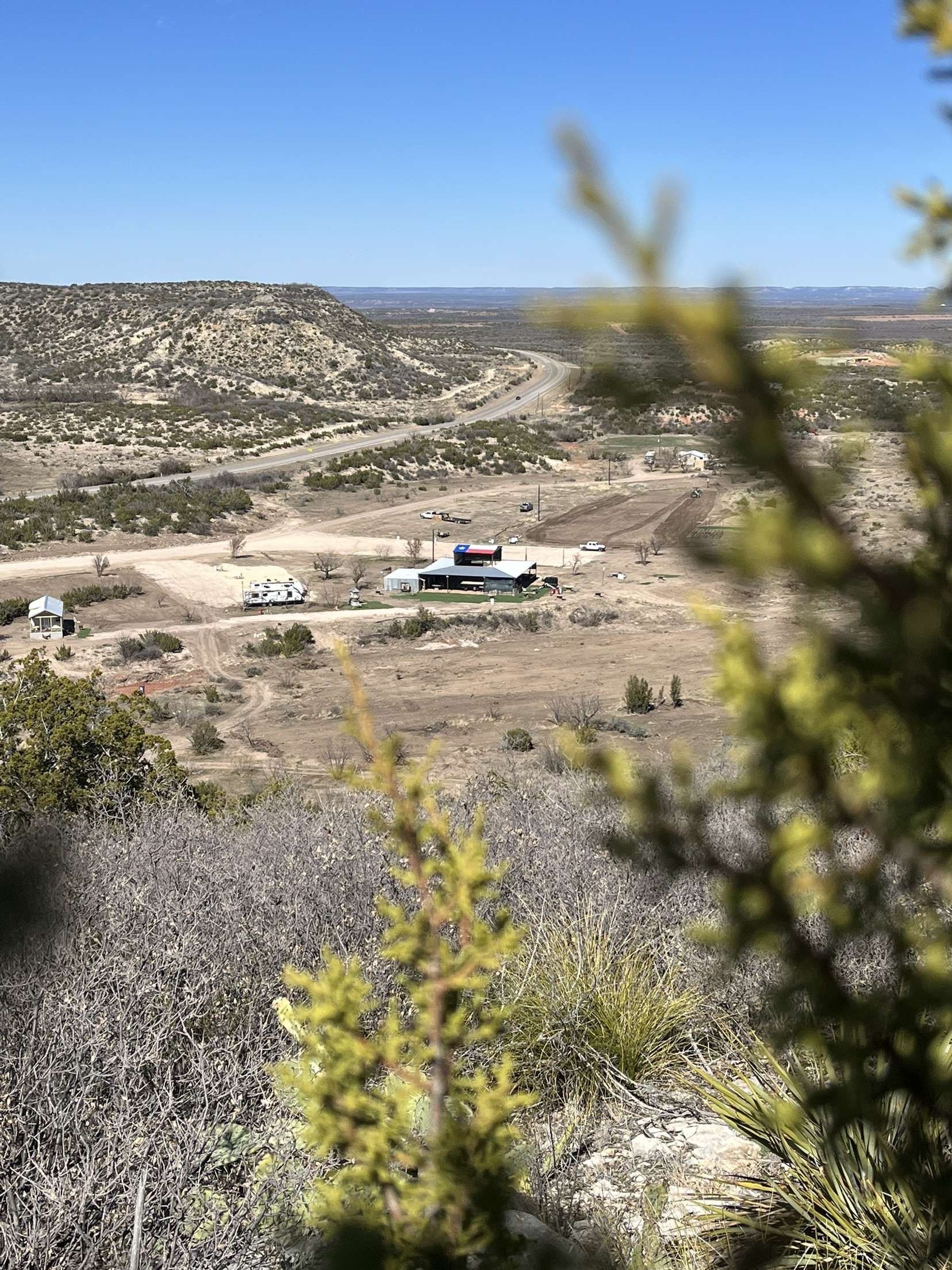 Arid hillside valley with scattered shrubs and a small white building under a clear blue sky