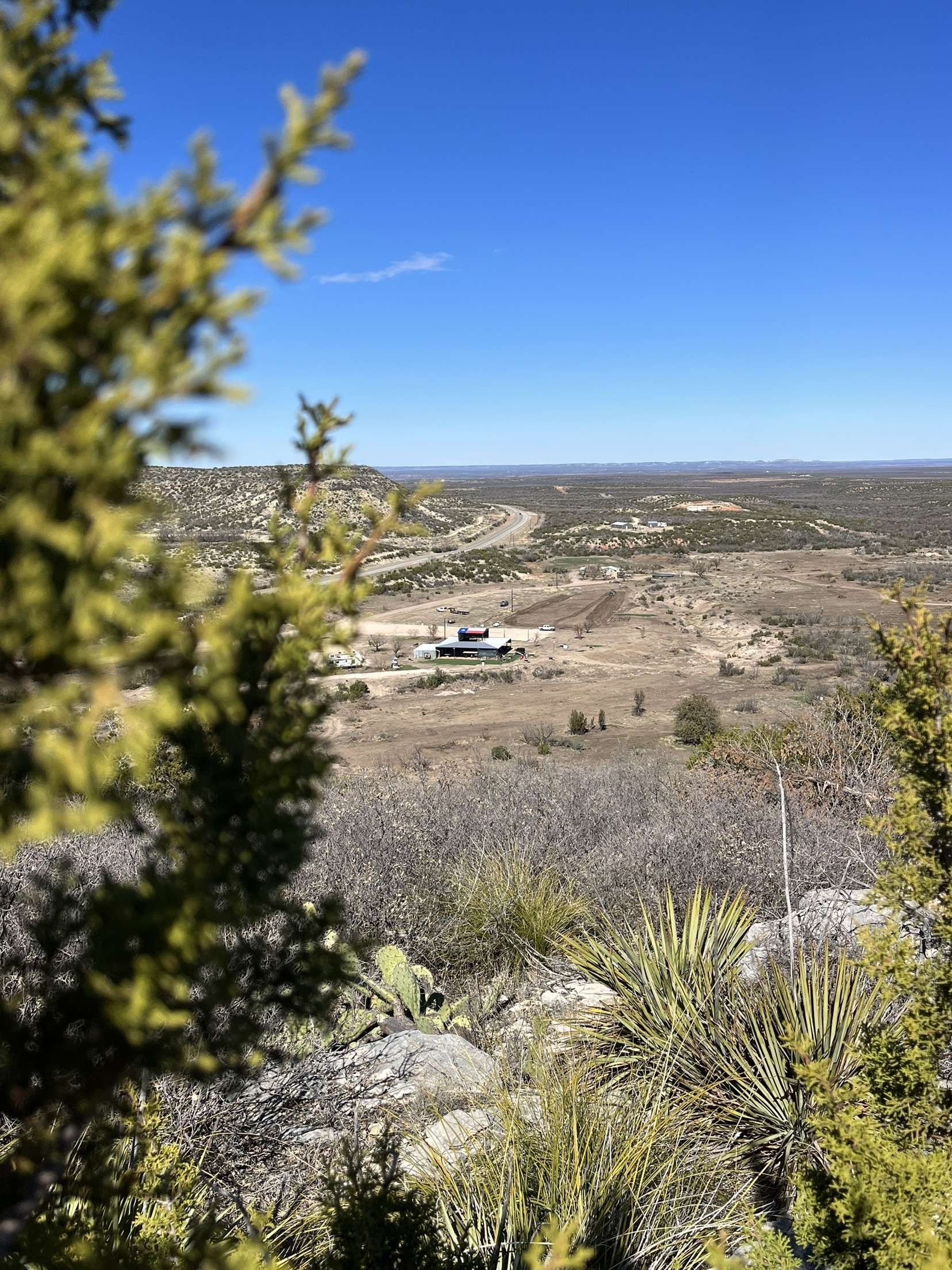 Rocky desert landscape with a distant building under a clear blue sky, framed by trees and shrubs