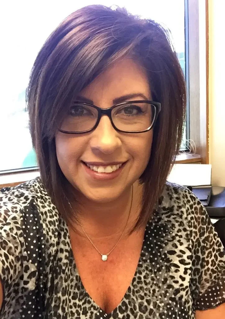 Woman with short dark hair, glasses, smiling, wearing patterned shirt, pearl necklace.
