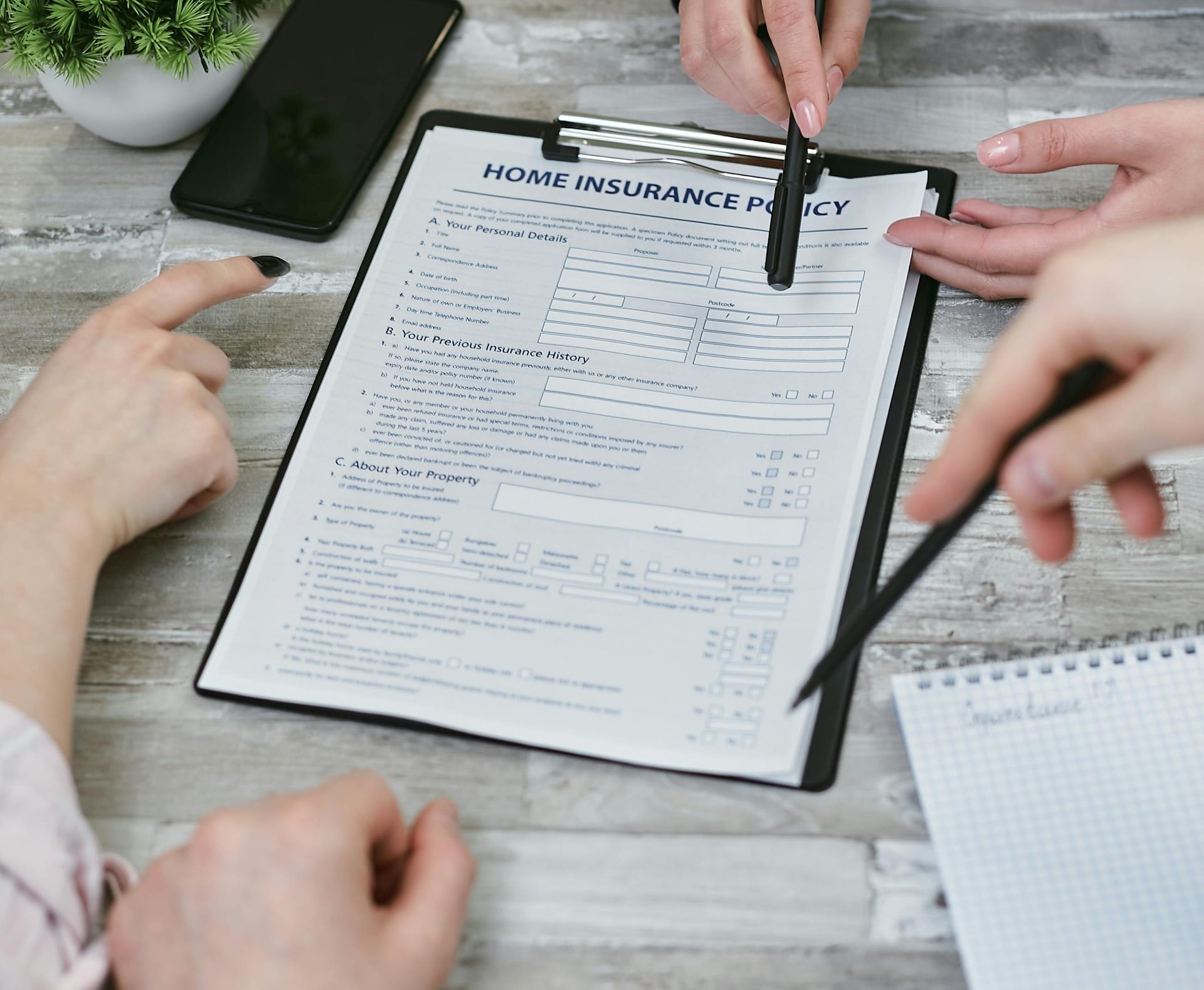People reviewing a home insurance policy. Hands pointing at the document, phone and notebook on a wooden table.