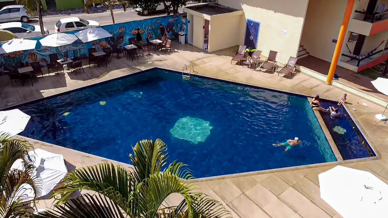 Piscina em formato de diamante com nadadores, rodeada por um pátio com mesas e guarda-sóis. É possível ver o edifício e as palmeiras.