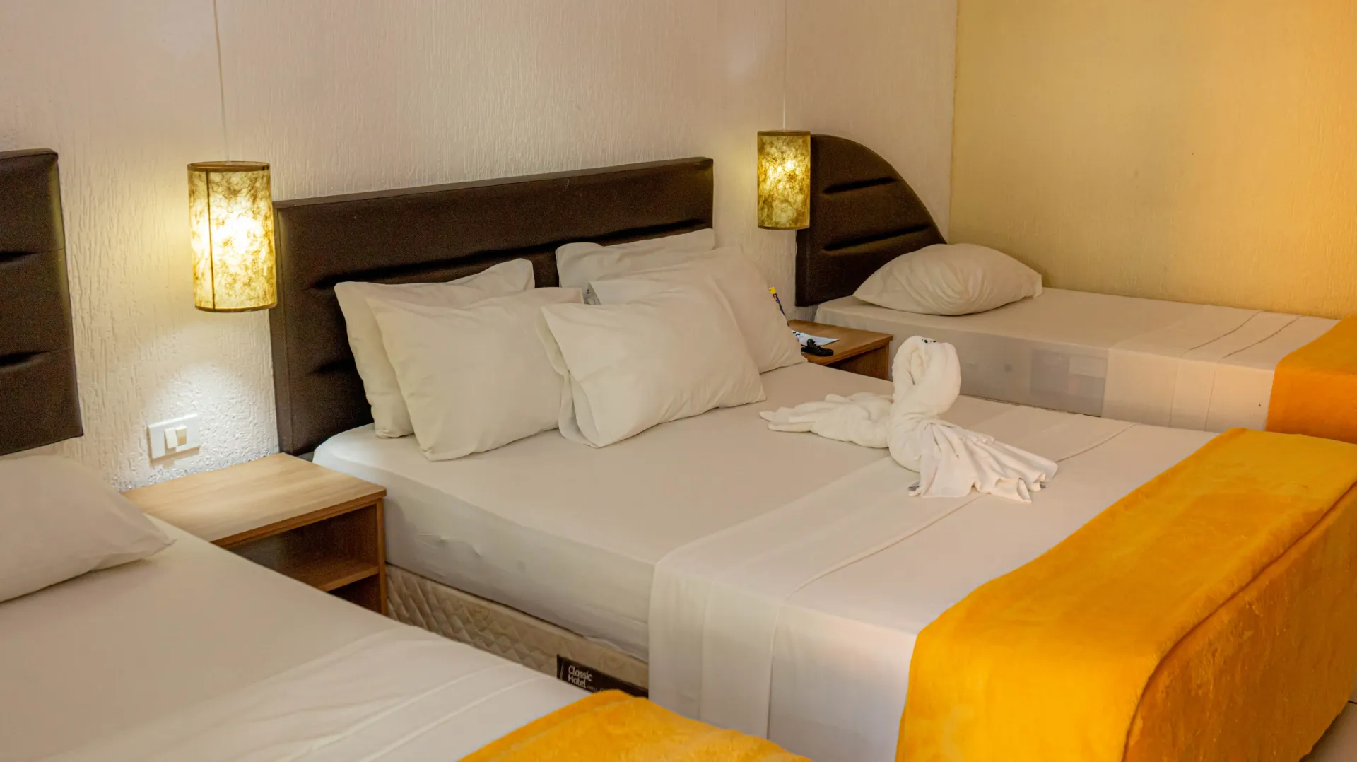 Quarto de hotel com três camas, lençóis brancos, detalhes em amarelo e abajures.