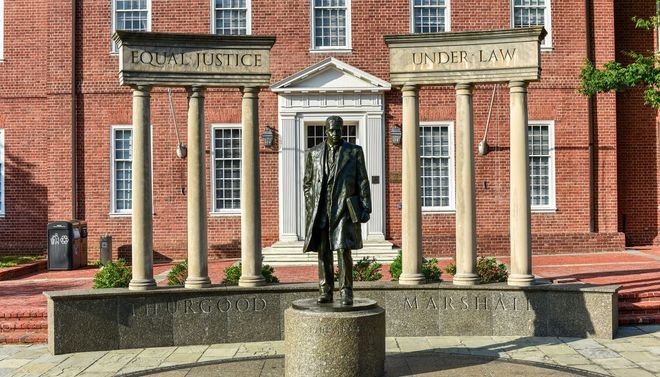 Statue before a red-brick courthouse with columns and signs reading “Equal Justice” and “Under Law”